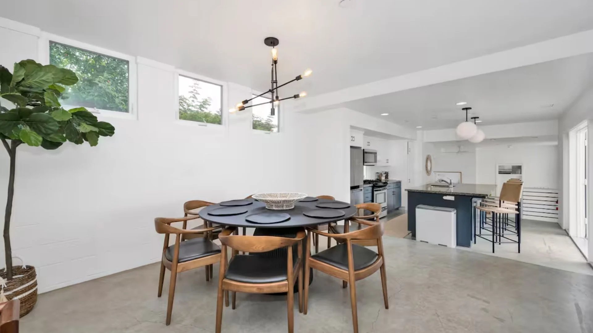 Modern open-concept kitchen and dining area with round wooden table, black placemats, six chairs, large windows, pendant and chandelier lighting, white walls, and a potted plant.