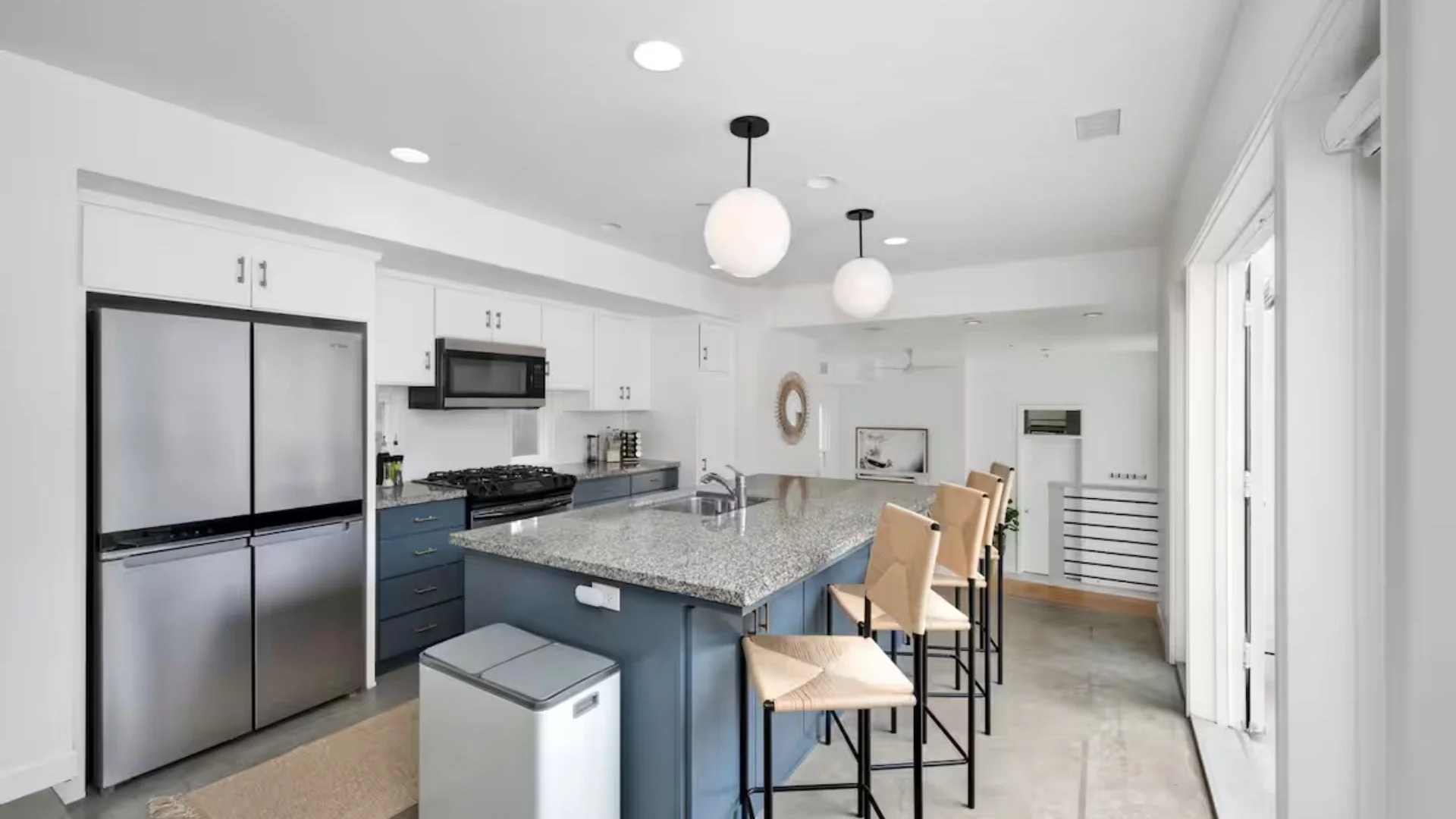 Modern kitchen with white cabinets, stainless steel refrigerator, black microwave, granite countertops, blue island with beige barstools, and pendant lighting.