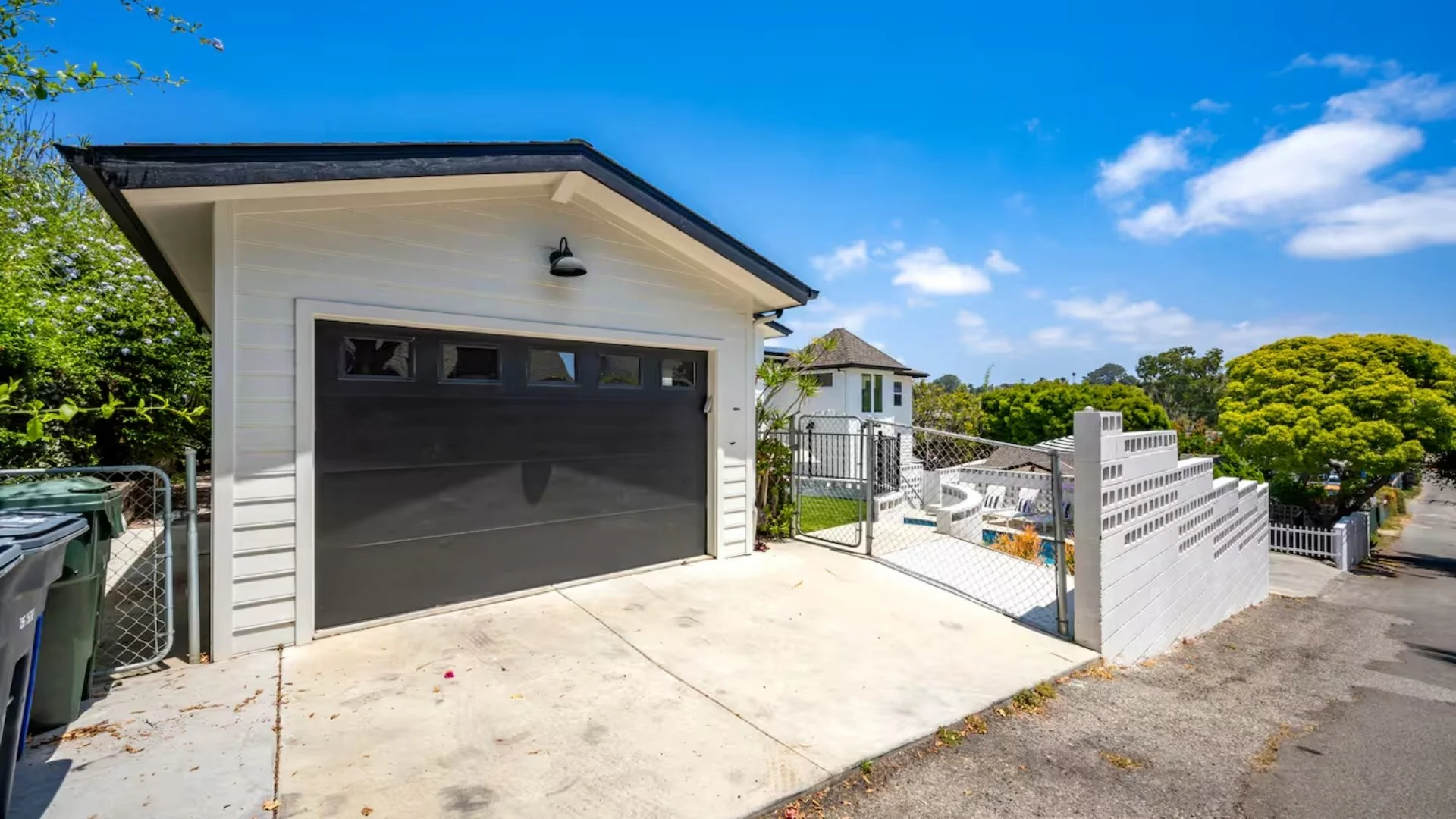 A residential garage with a black door, white siding, and a sloped roof. There are two trash bins on the left and a chain-link fence with a gate on the right, leading to a fenced backyard with a small patio and a white house in the background. Tall g