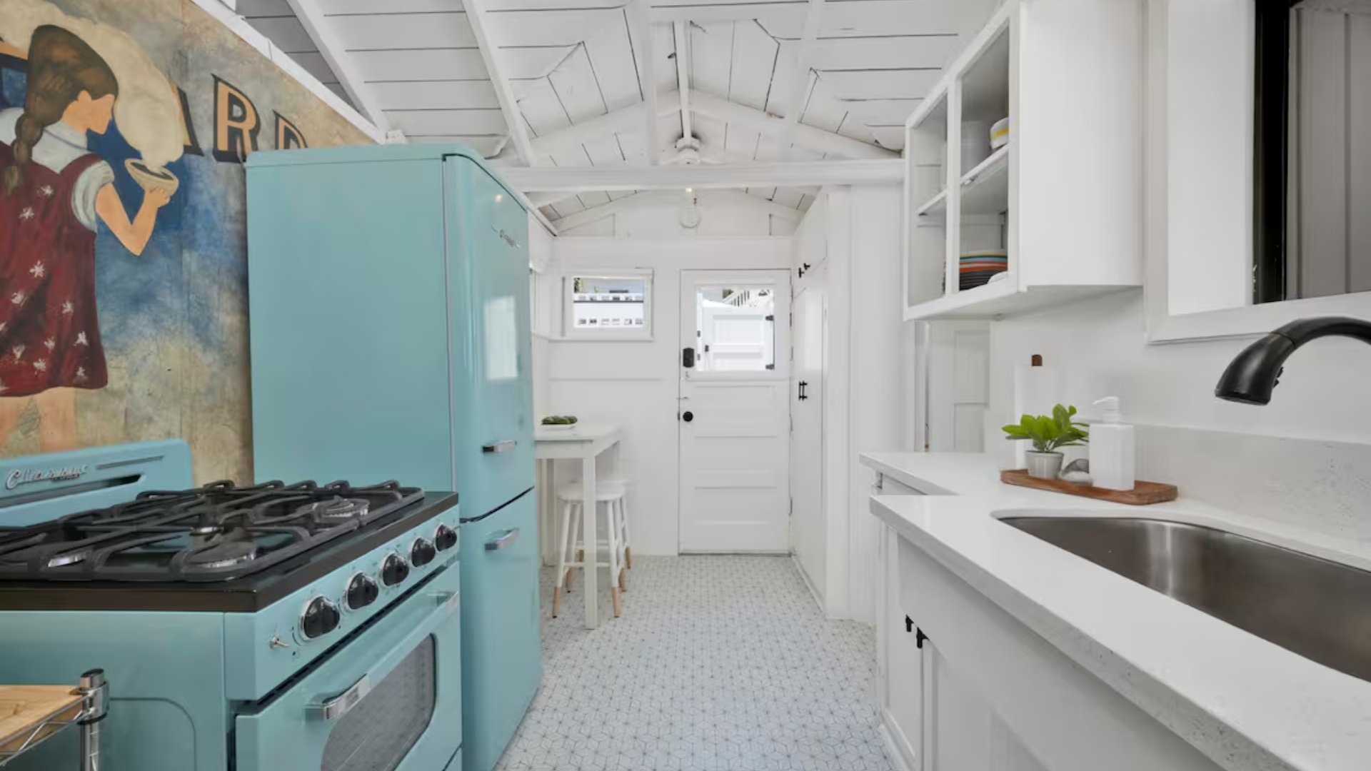 Bright white kitchen with a vintage blue stove and matching refrigerator, white cabinetry, a small counter with a plant, and a painted mural of a girl holding a bowl on the wall.