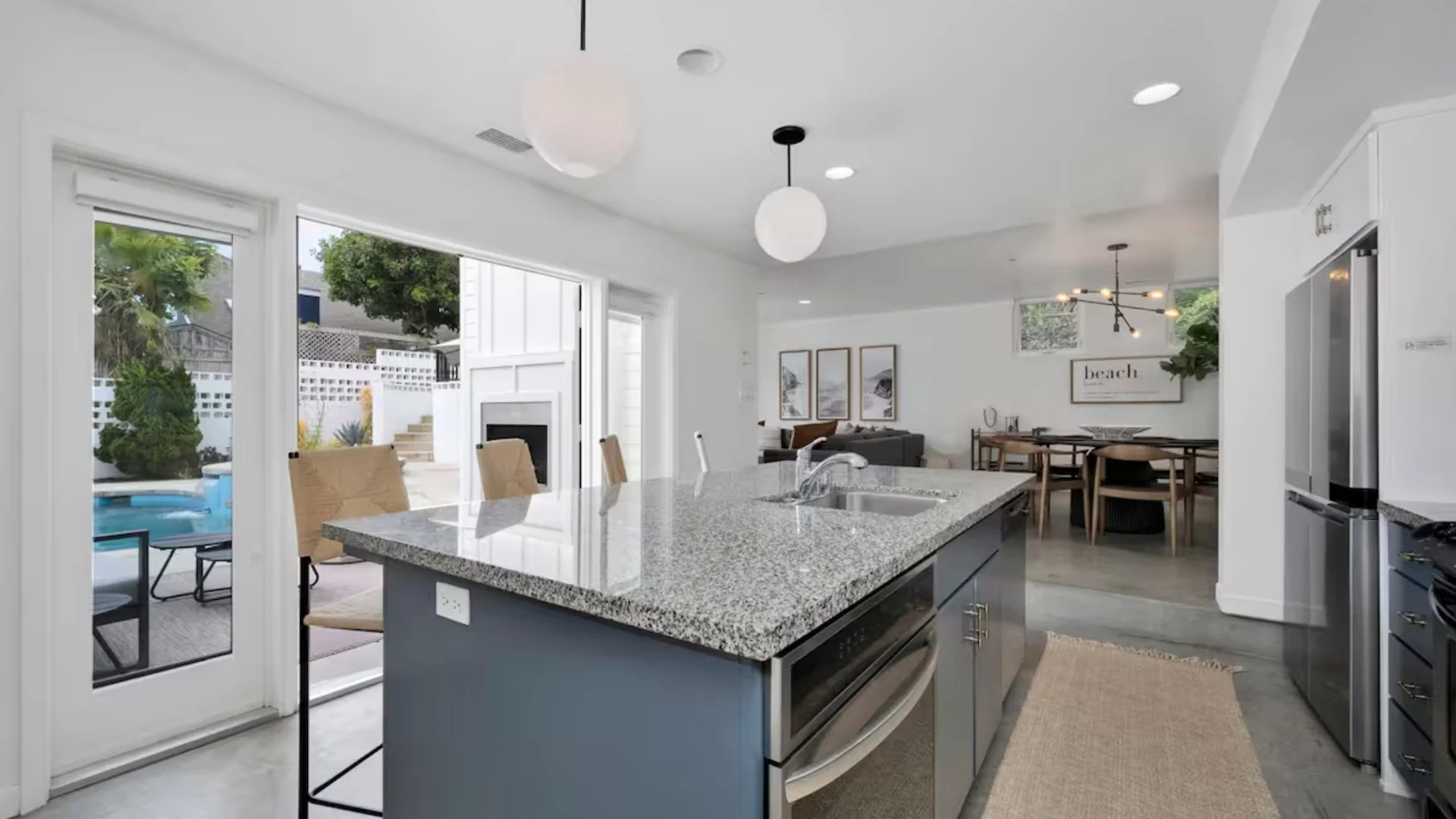 Modern open-concept kitchen and living space with a granite kitchen island, stainless steel appliances, dining area, and outdoor patio with pool and greenery visible through sliding glass doors.