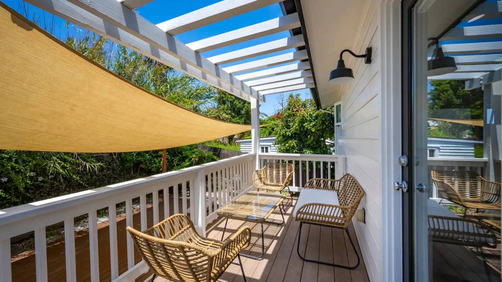 Small balcony with wicker chairs and table, partial shade sail, white railing and siding, black wall-mounted lights, surrounded by green trees and shrubs.