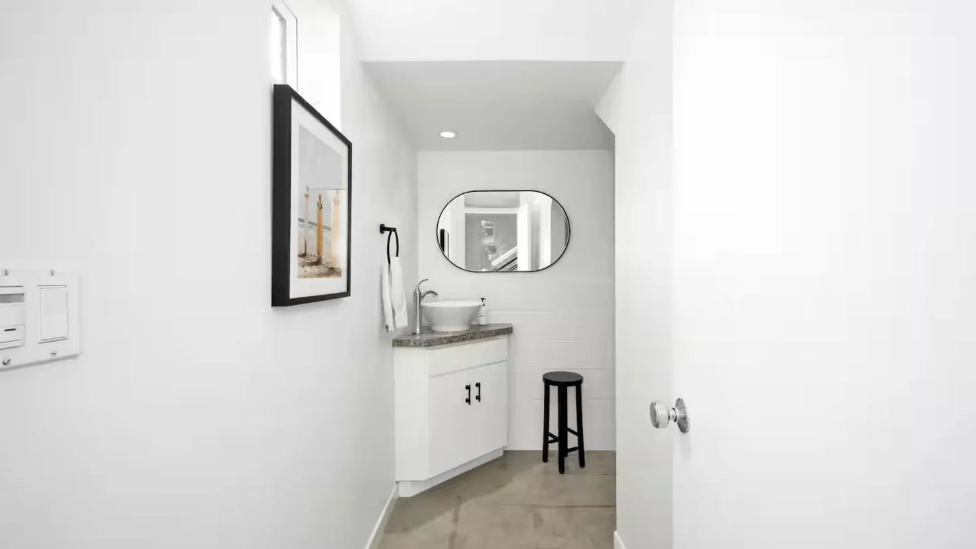 A small bathroom with white walls, a cabinet with a granite countertop, a vessel sink, a round mirror, a black stool, and wall art. There's a small window near the ceiling, and the door is partially open.