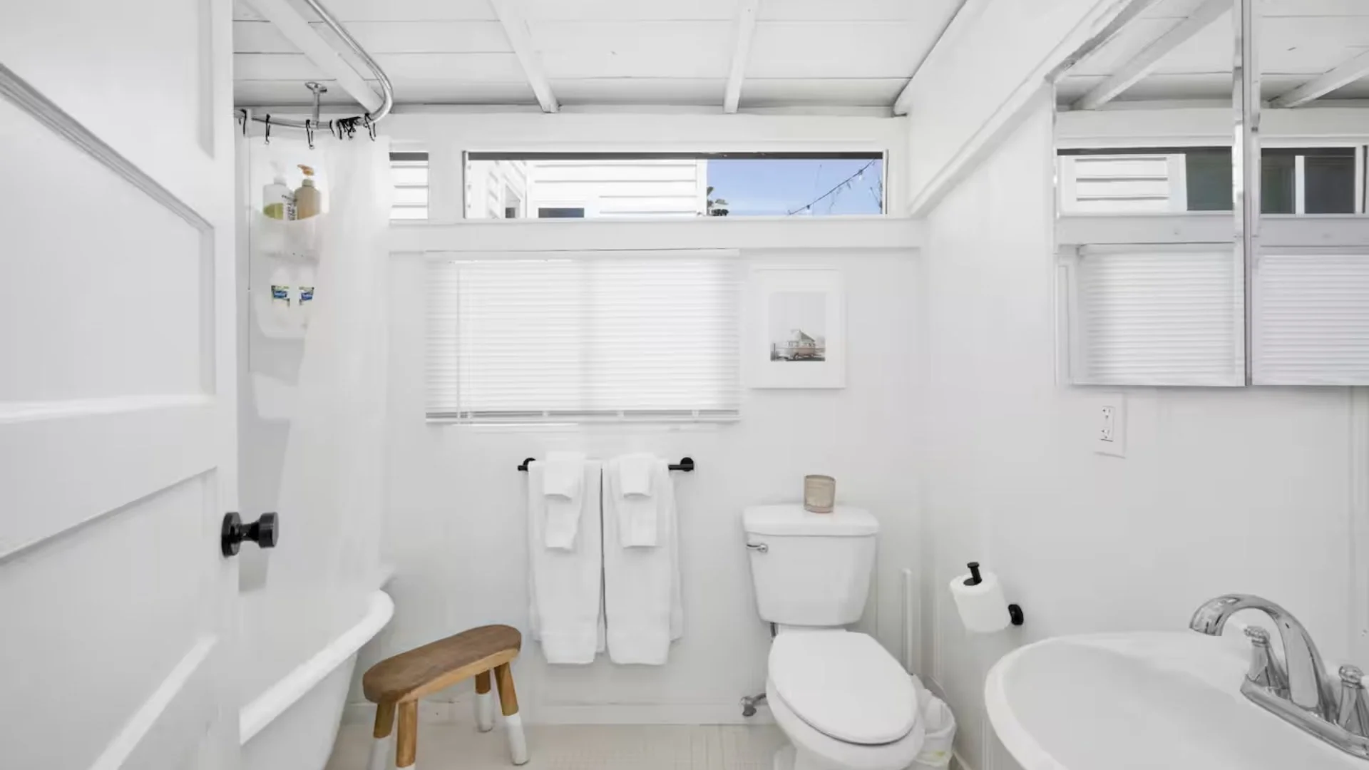 Bright white bathroom with window shades, white towels on a black towel rack, toilet with a small candle on top, and a white sink with chrome faucet, with windows showing a blue sky and neighboring houses.