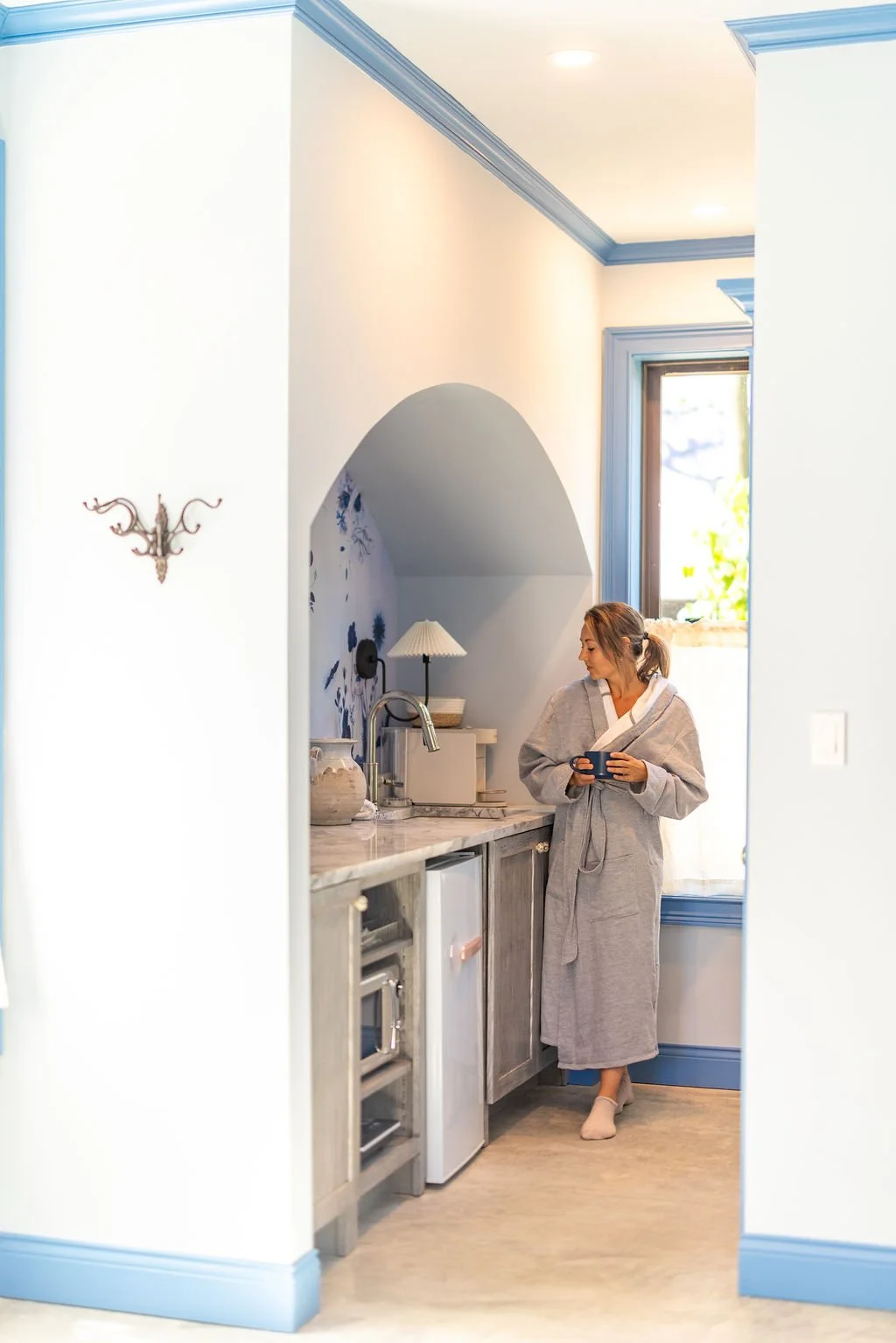 A woman in a bathrobe holding a mug, standing in a cozy kitchen with a marble counter, a small refrigerator, a sink, a lamp, and decorative vases. She appears to be enjoying a relaxing morning.