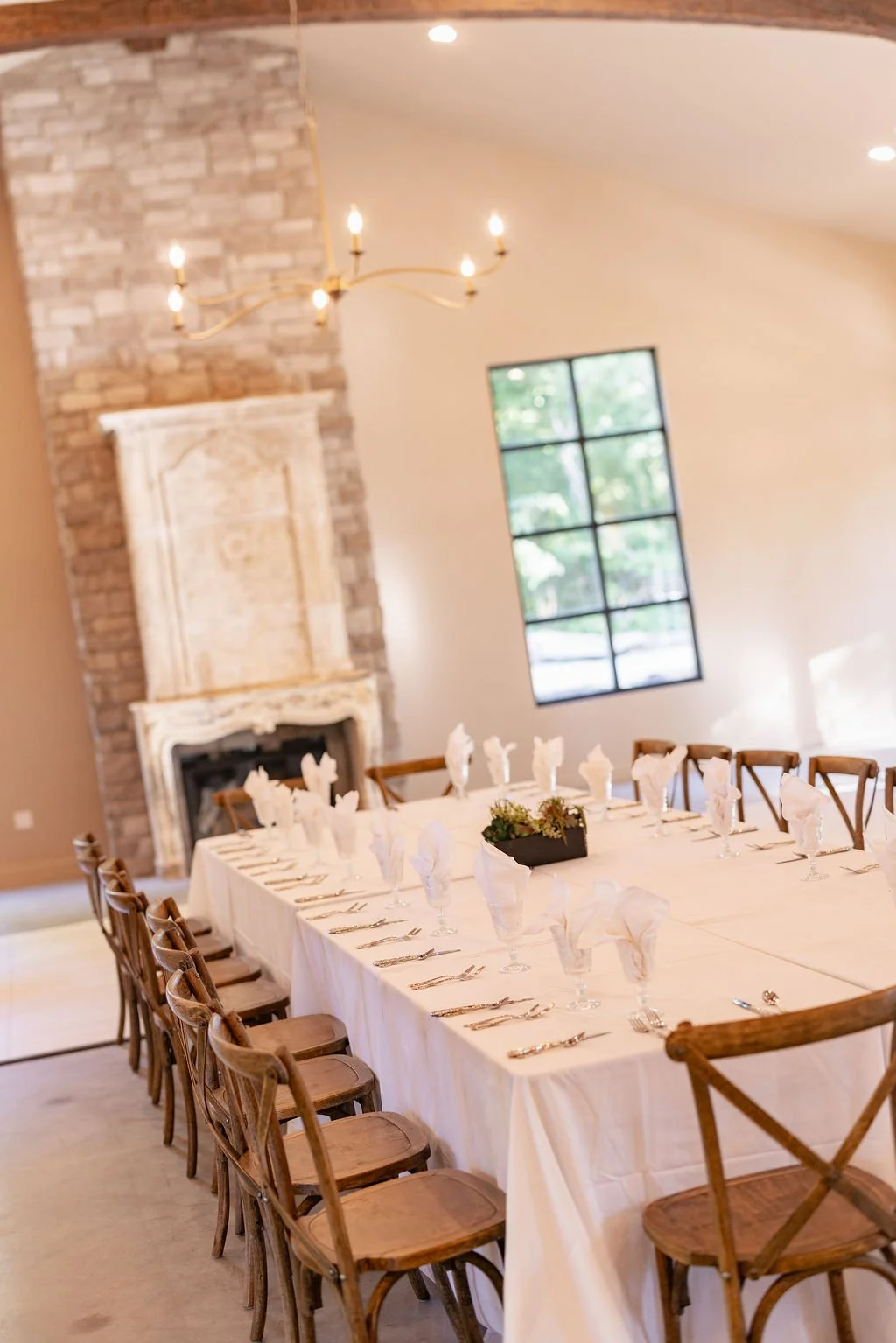 A dining table set for a formal event in a room with a stone fireplace and large window, featuring white tablecloth, napkins, and silverware.