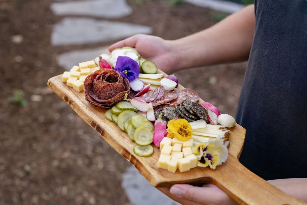 Person holding a wooden platter with assorted cheeses, cured meats, pickles, and edible flowers.