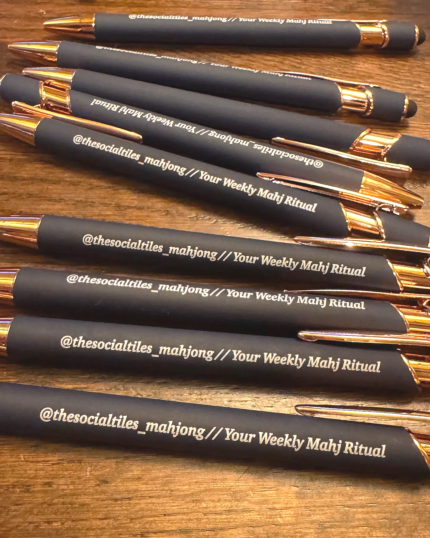 A group of black pens with gold accents and white text that reads '@thesocialties_mahjong // Your Weekly Mahj Ritual' are laying on a wooden surface.