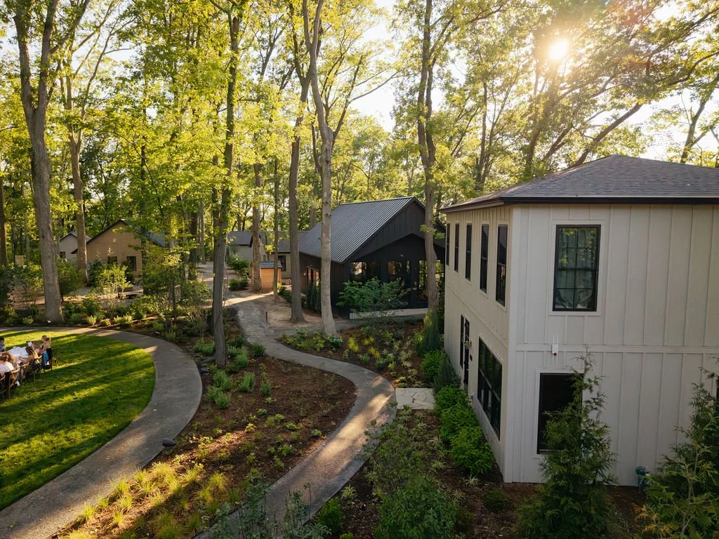 A peaceful outdoor setting with a well-maintained lawn, winding pathways, and several houses surrounded by tall trees under a sunny sky.