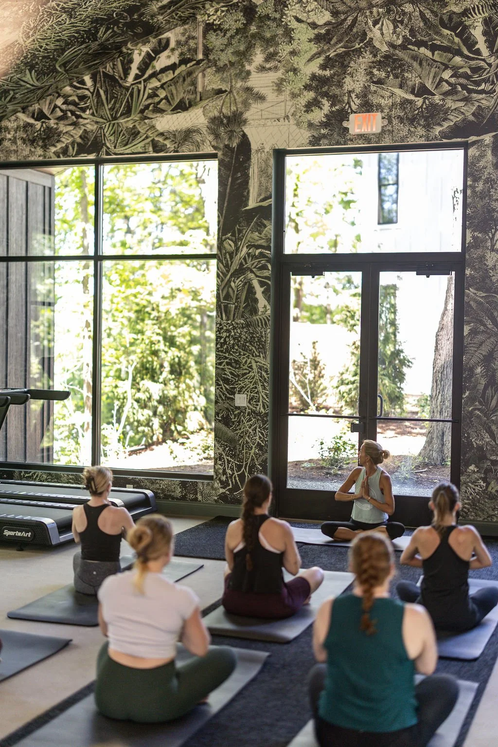 A yoga class taking place inside a room with large windows and lush outdoor scenery. Participants are seated on yoga mats, facing a woman instructor who is sitting cross-legged in a meditative pose.