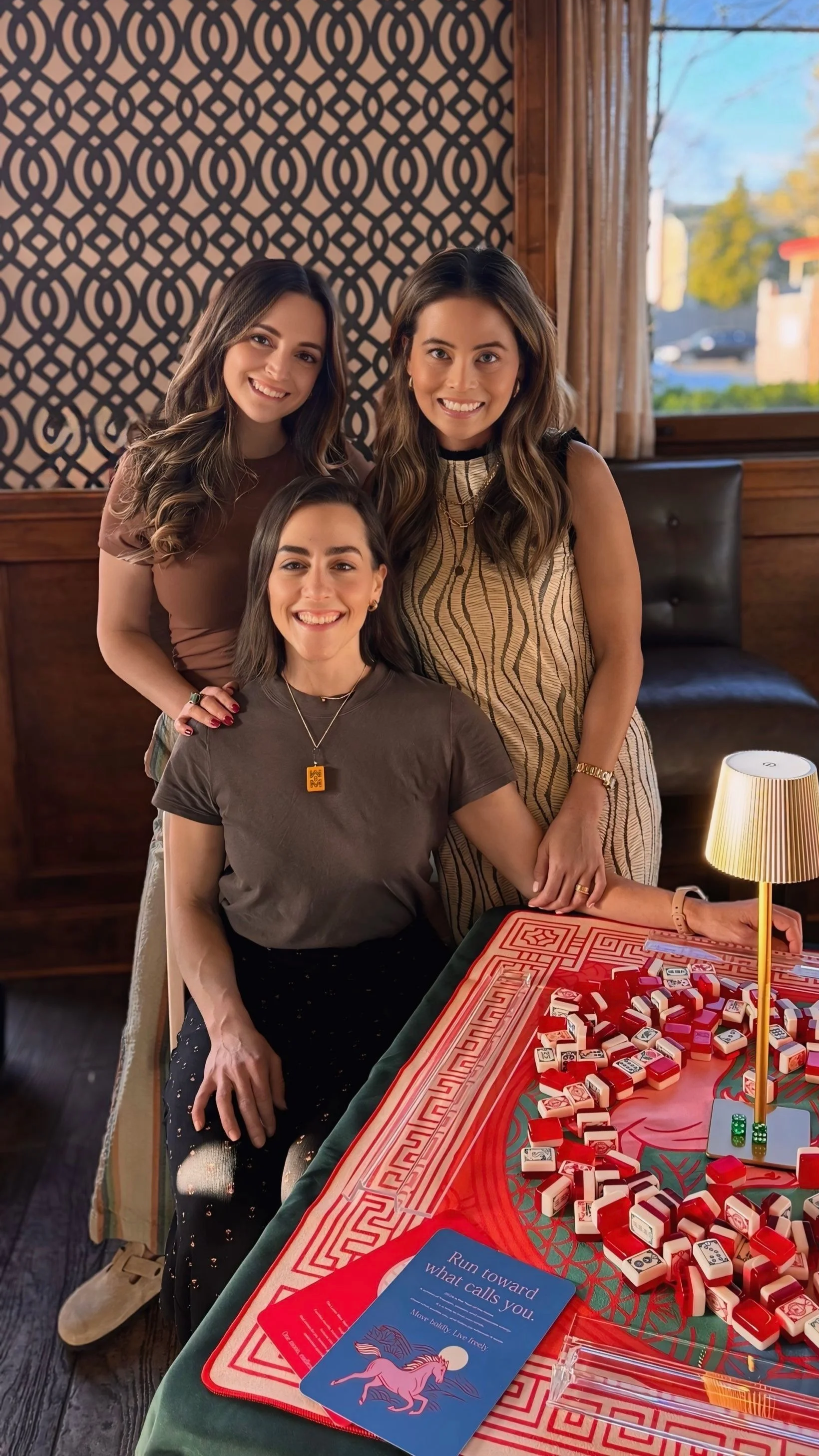 Three women smiling around a table with Mahjong tiles. Learn how to play mahjong with them in Glenview, Illinois