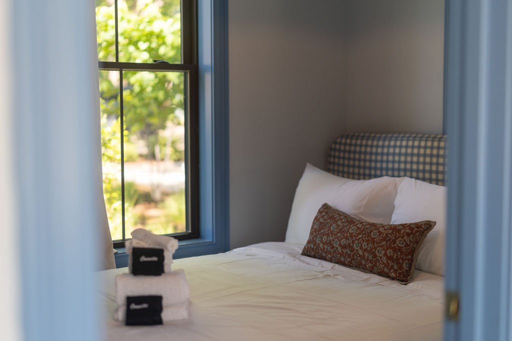 A cozy bedroom with a neatly made bed, pillow, and decorative pillow, next to a window showing greenery outside.