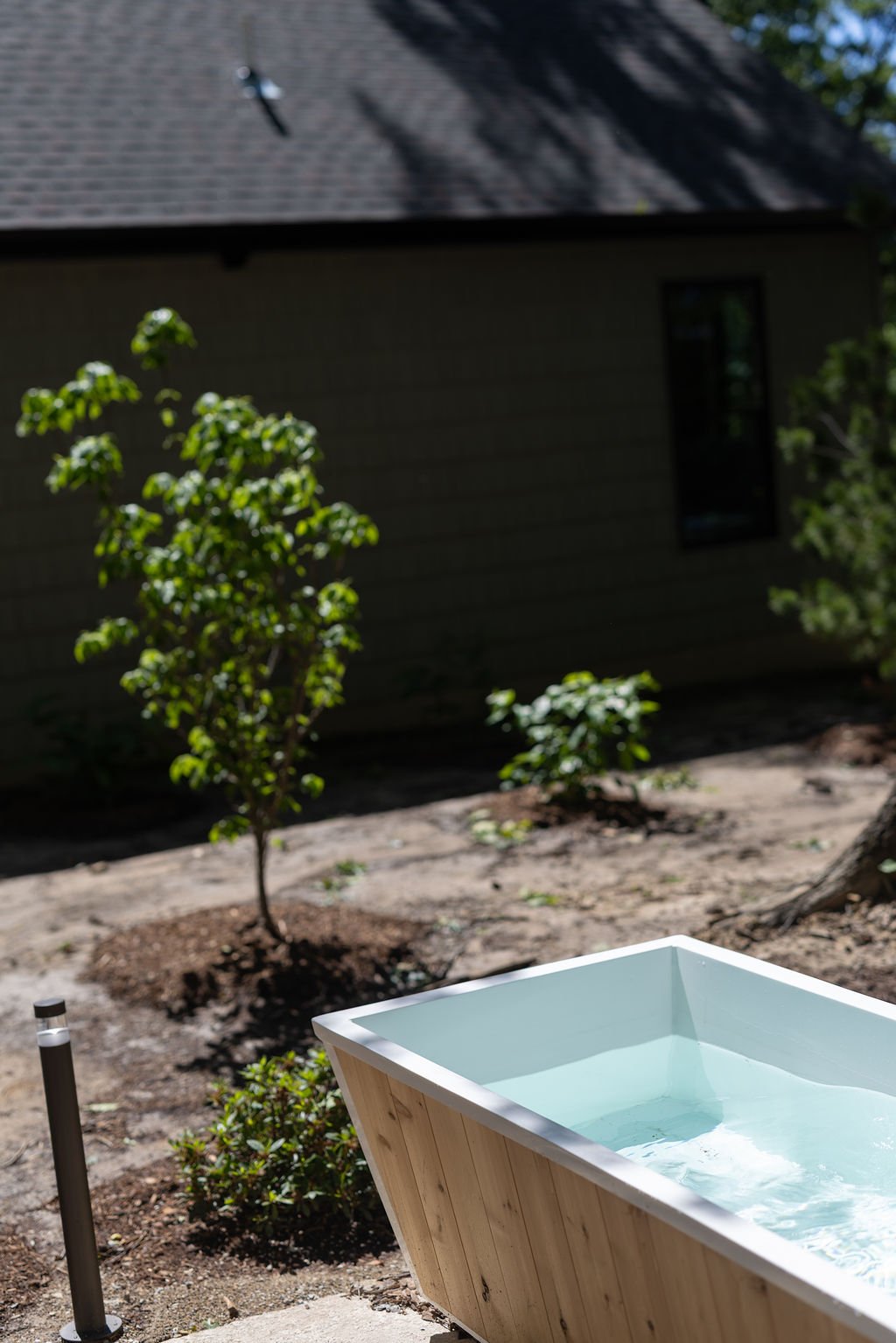 A small wooden hot tub filled with water outdoors, in a yard with small green trees and bushes, near a house with dark gray siding and a window.