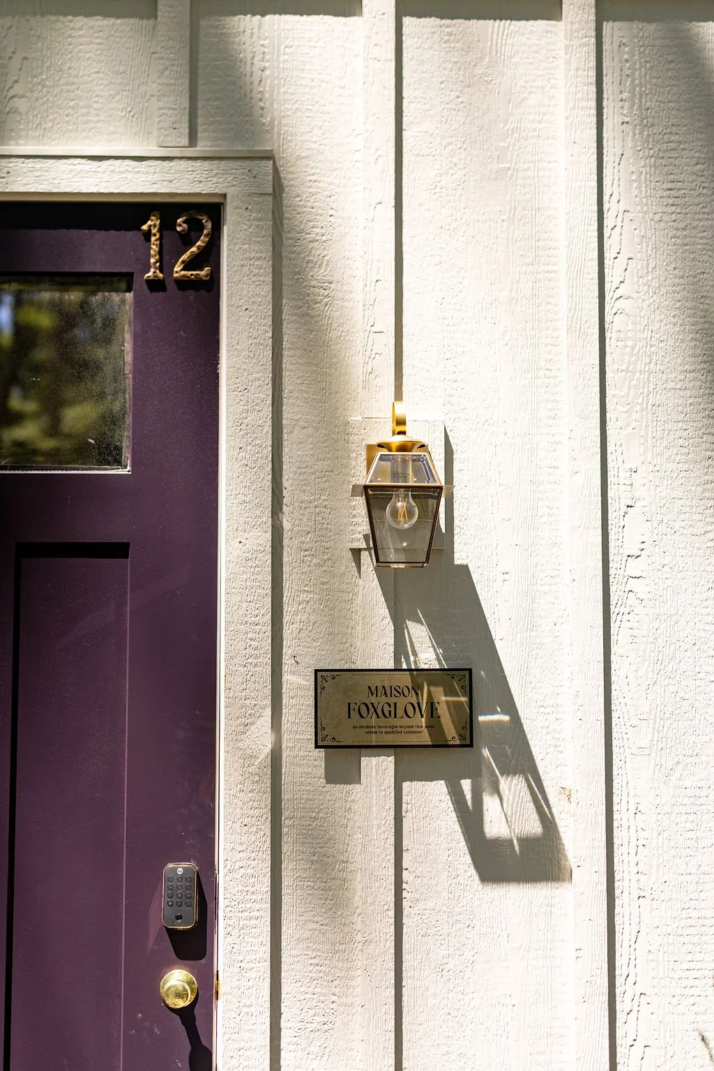 Close-up of a house exterior showing a purple door with the number 12, a lantern-style wall light, and a sign reading 'Maison Foxglove' on a white wooden wall.