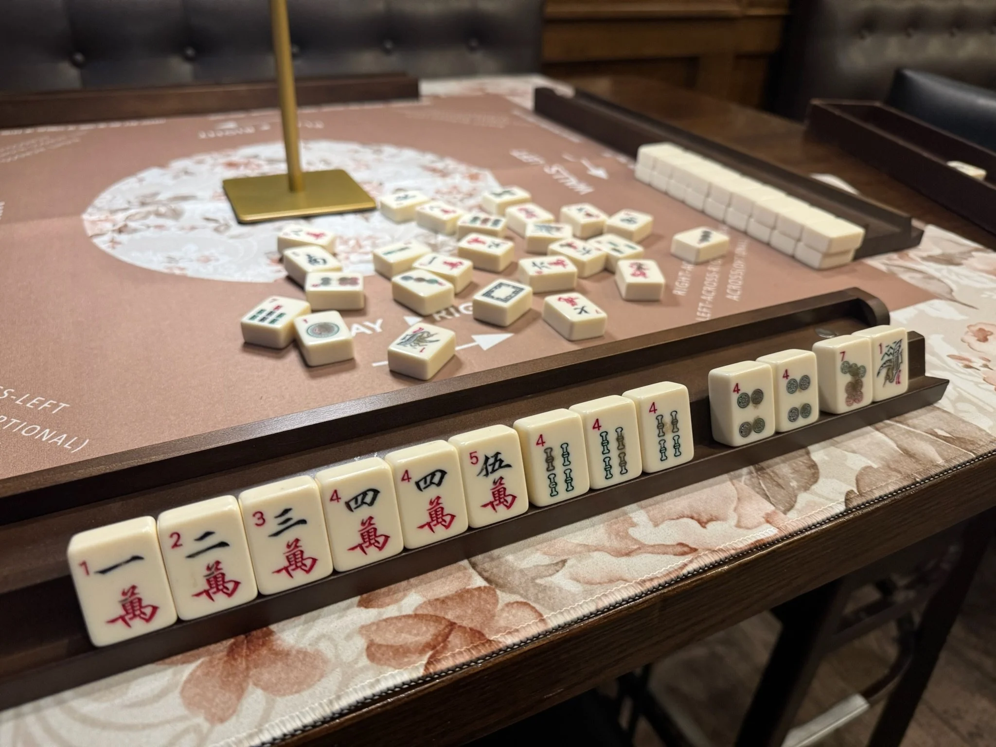Close-up of a Mahjong game in progress on a table with tiles arranged in a row and others stacked, with a small brass stand.