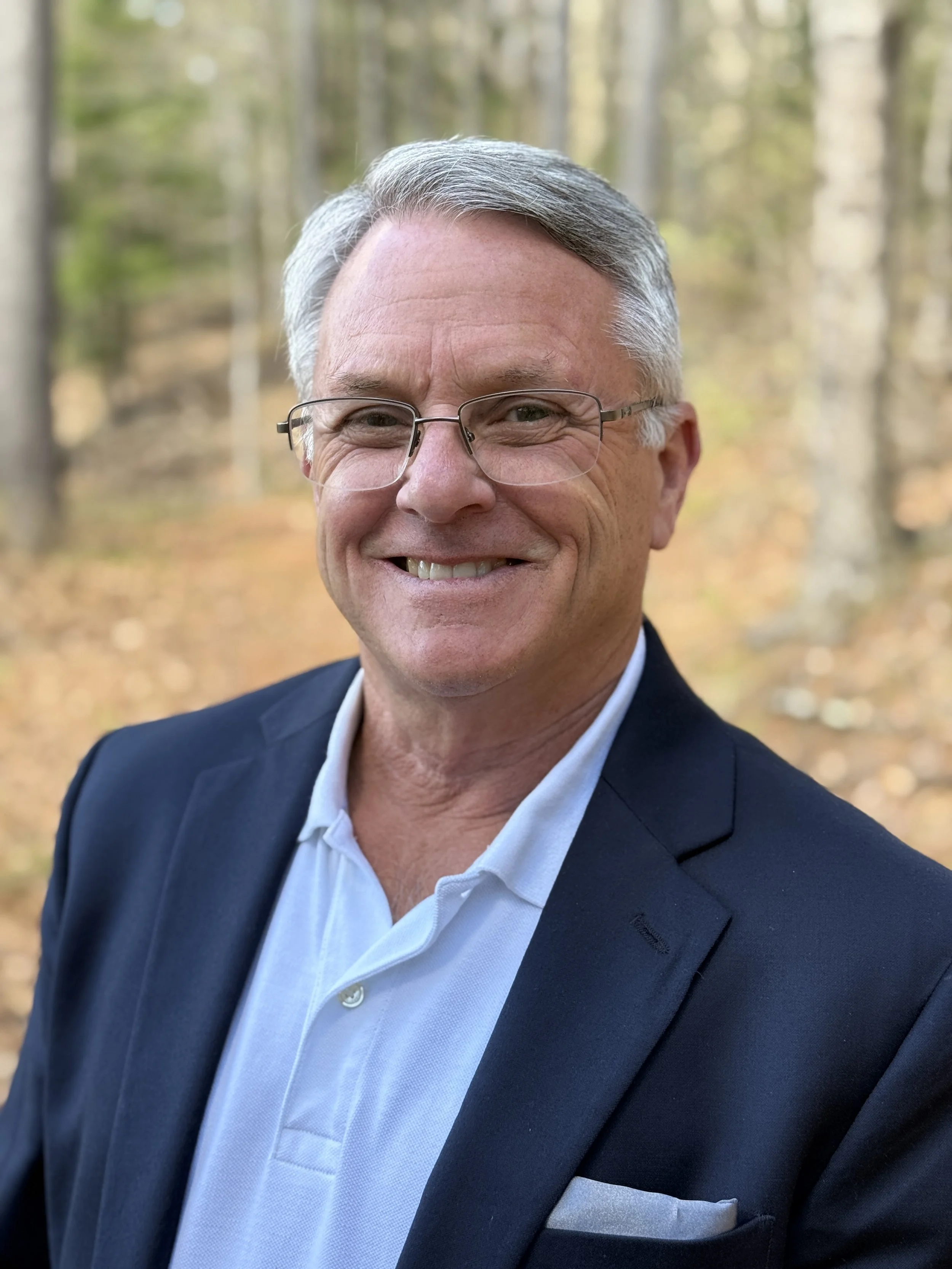 A smiling middle-aged man with gray hair and glasses, dressed in a navy blazer and white shirt, standing outdoors in a wooded area with trees and fallen leaves.