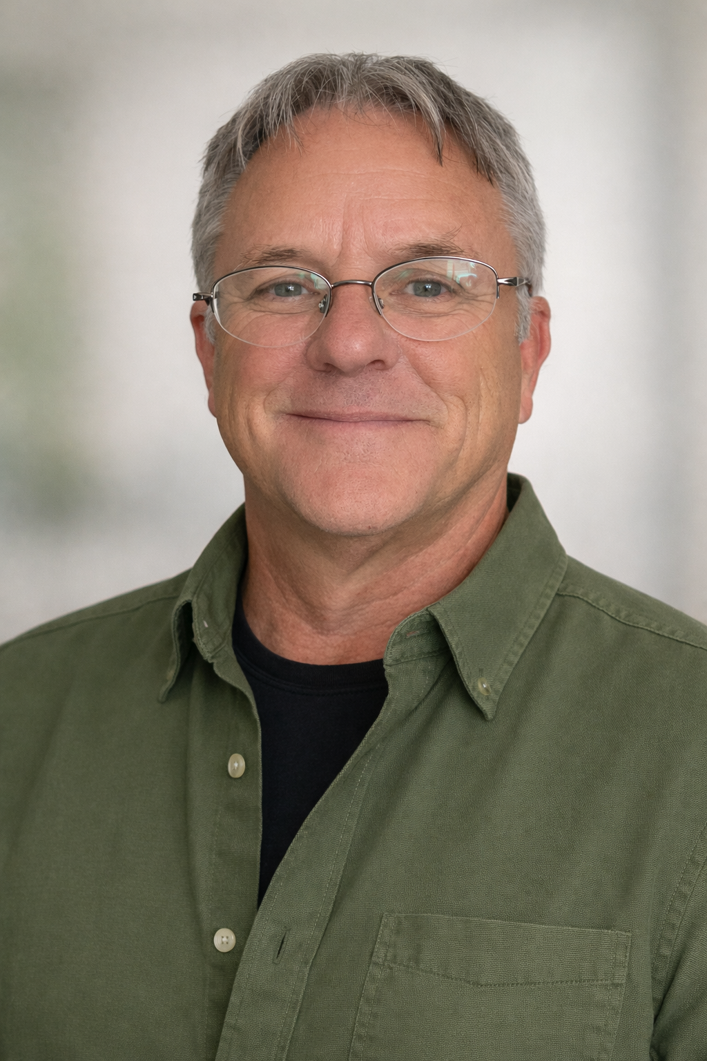 A middle-aged man with gray hair, glasses, and light skin, smiling, wearing a green button-up shirt over a black T-shirt, standing against a neutral background.