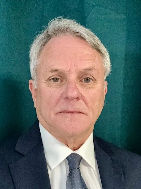 Portrait of an older man with gray hair wearing a suit, white shirt, and a patterned tie, standing in front of a teal-colored background.