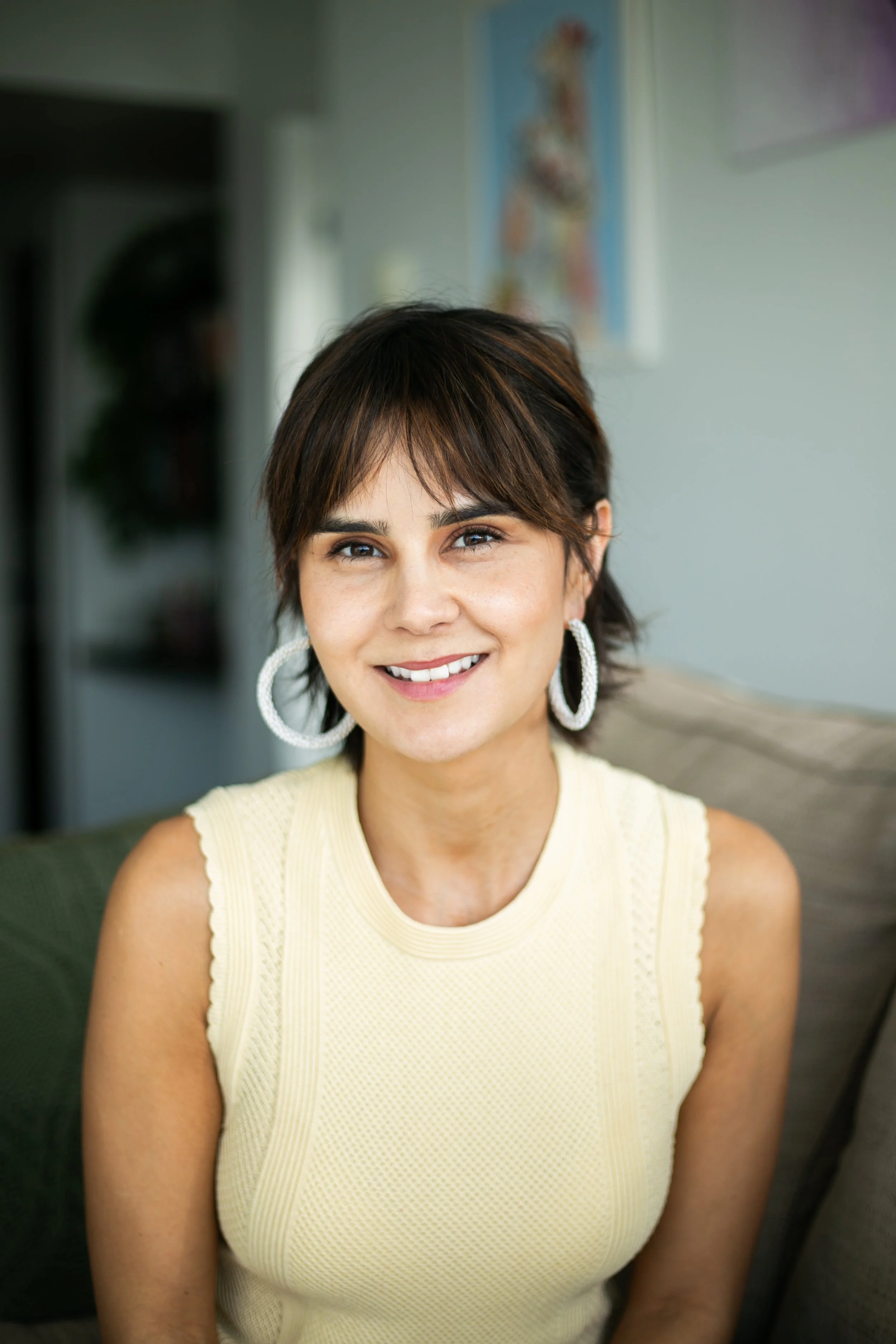 A woman with short brown hair and large white hoop earrings smiling while sitting indoors, wearing a sleeveless light yellow top.