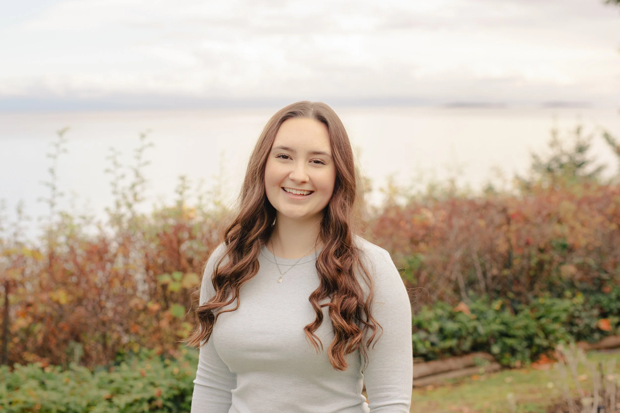 A young woman with long wavy brown hair smiling outdoors in front of colorful autumn shrubbery and trees during daytime.