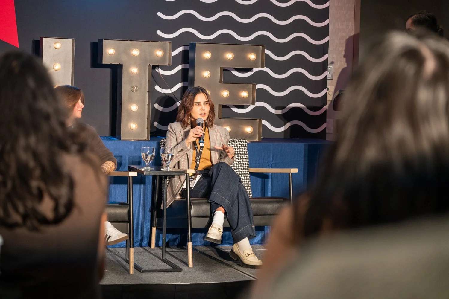 Woman speaking into a microphone on a stage with illuminated letters spelling 'TE' behind her, in front of an audience at a panel or conference.