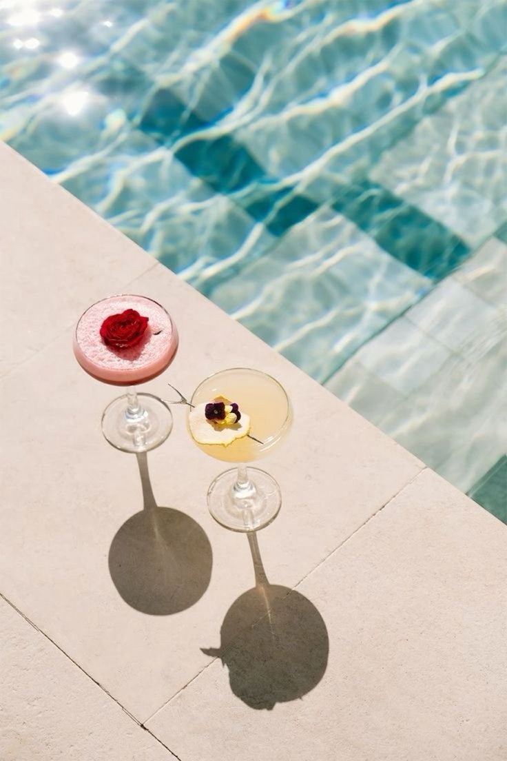 Two cocktails with fruit garnishes on a poolside table, with the pool water visible in the background.