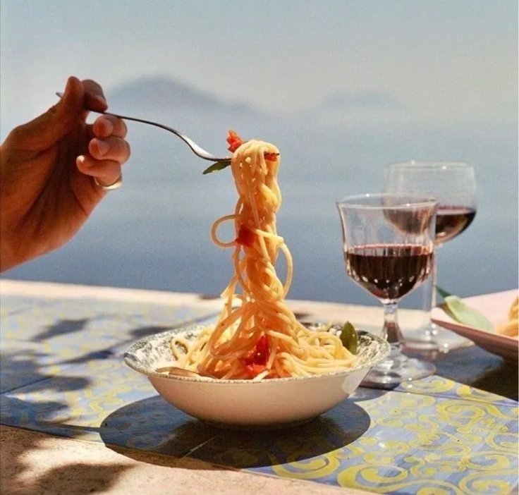A bowl of spaghetti with a fork holding a serving of noodles, set on an outdoor table with a scenic water view. There are two glasses of red wine next to the bowl.