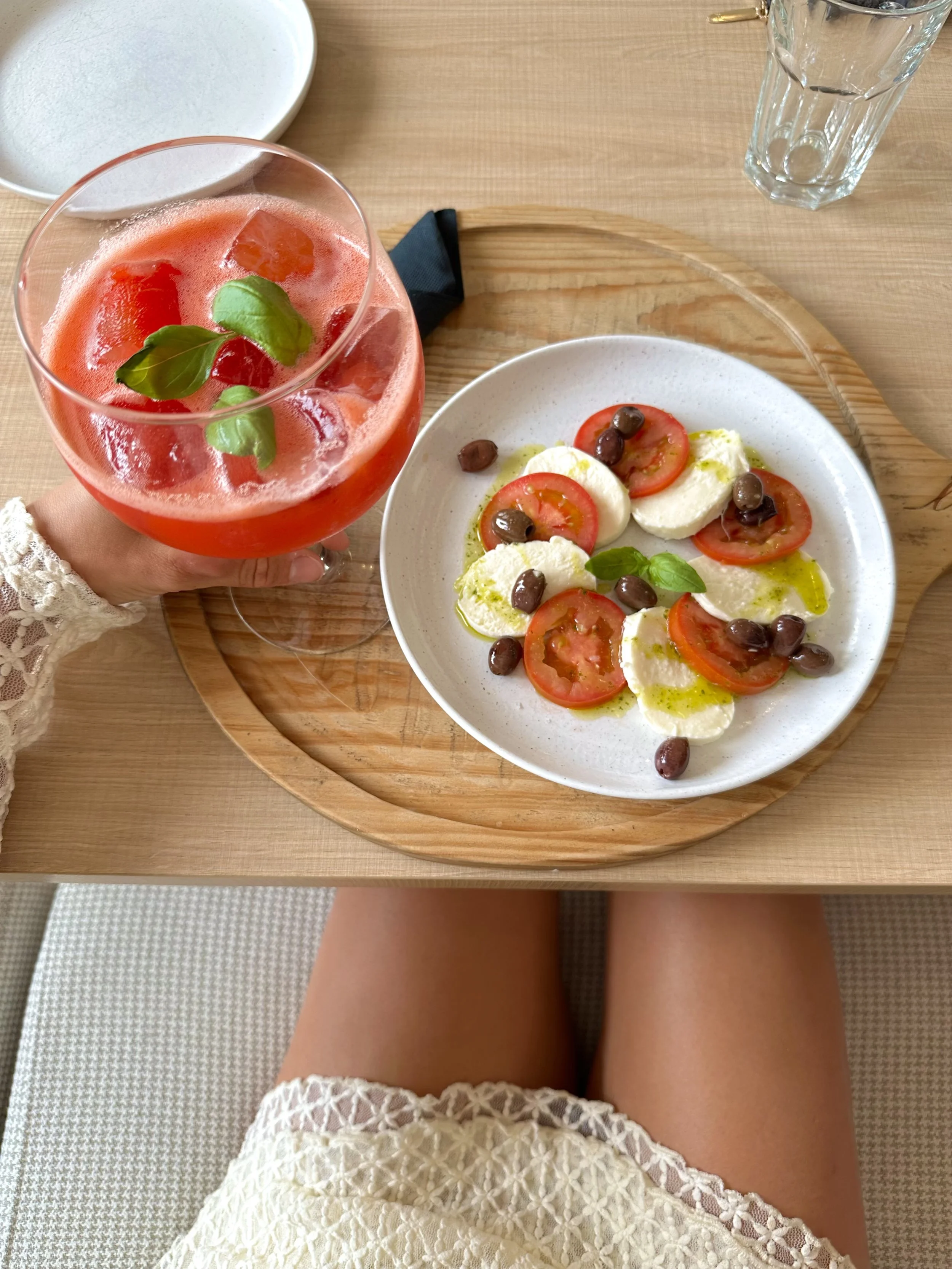 A plate with a Caprese salad consisting of sliced tomatoes, mozzarella cheese, green basil leaves, and black olives on a wooden tray.