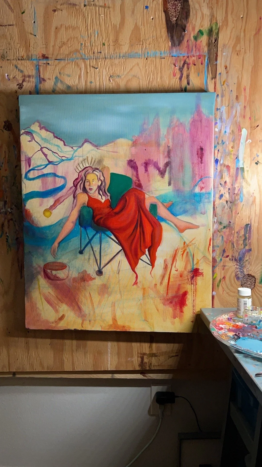 Painting of a woman in a red dress reclining in a green chair, with a colorful abstract background, on a canvas against a splattered paint wall.