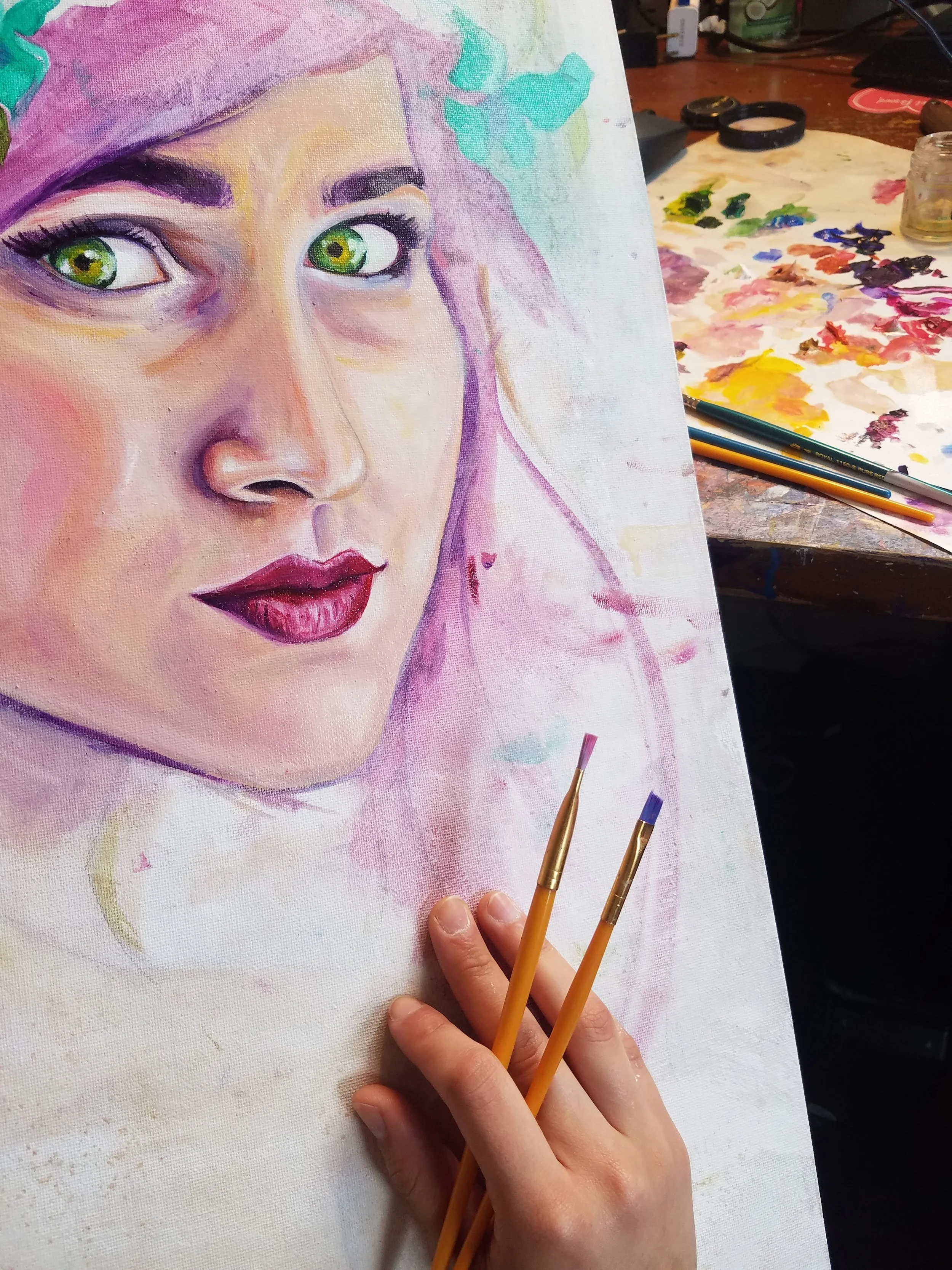 A partially completed watercolor painting of a woman's face on canvas, with vibrant green eyes, pink lips, and purple and pink hair, sitting on a cluttered table with painting supplies and brushes.