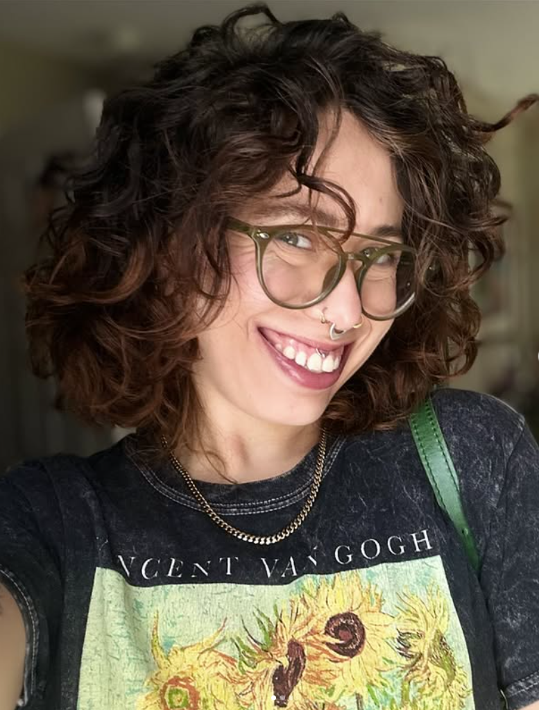 A young woman with curly brown hair, glasses, and a septum piercing, smiling and wearing a Vincent van Gogh T-shirt with sunflowers, a gold chain necklace, and a green strap over her shoulder.