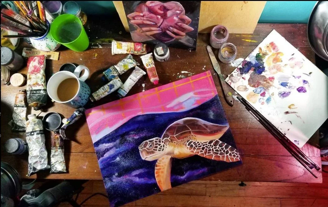 An artist's workspace with a painting of a sea turtle swimming through water and a sunset sky. The desk has various paint tubes, brushes, a cup of coffee, and a palette with mixed colors.