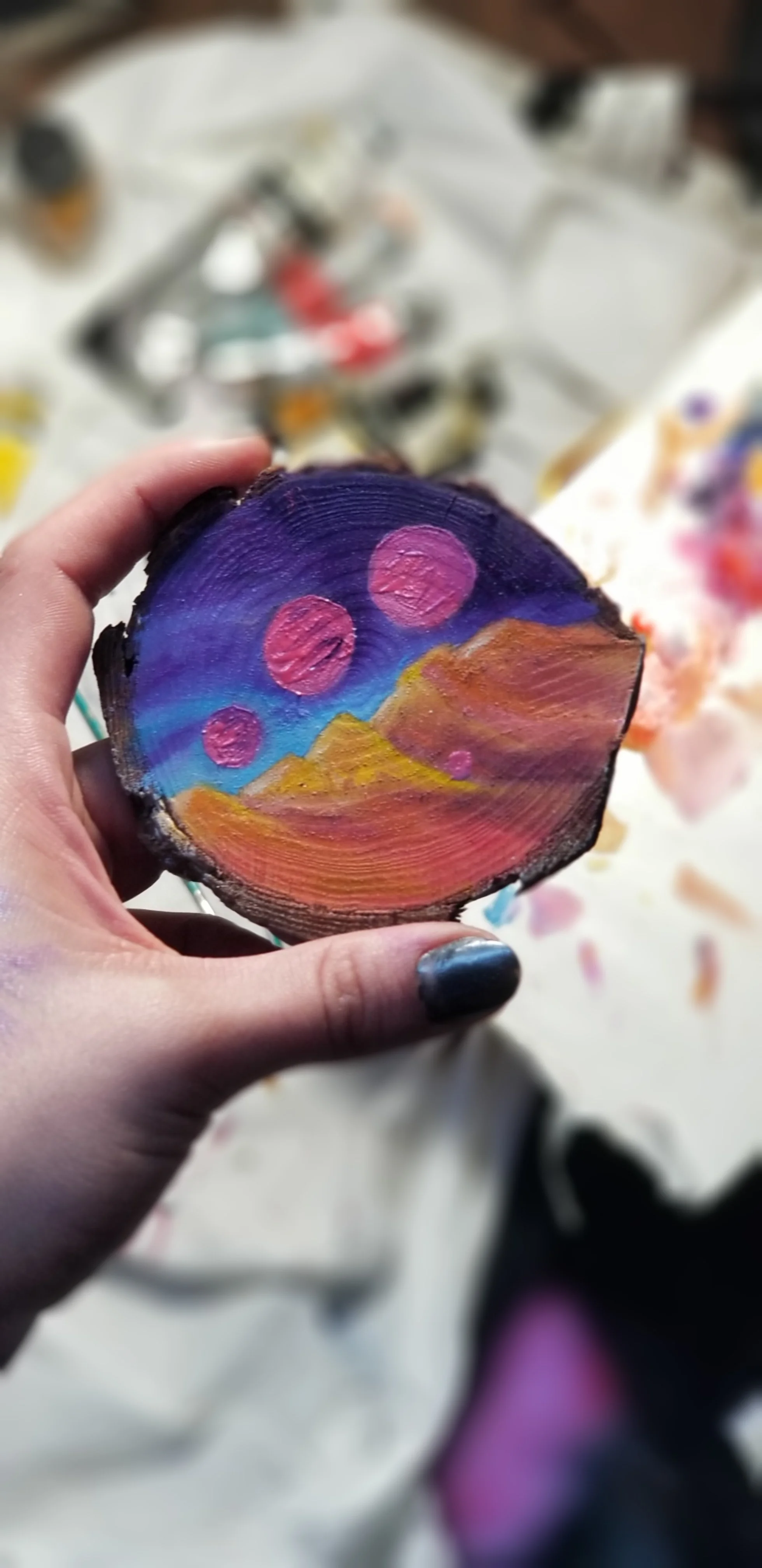 A hand holding a painted wooden slice depicting a colorful landscape with mountains, a purple sky, and pink circles representing planets or moons.