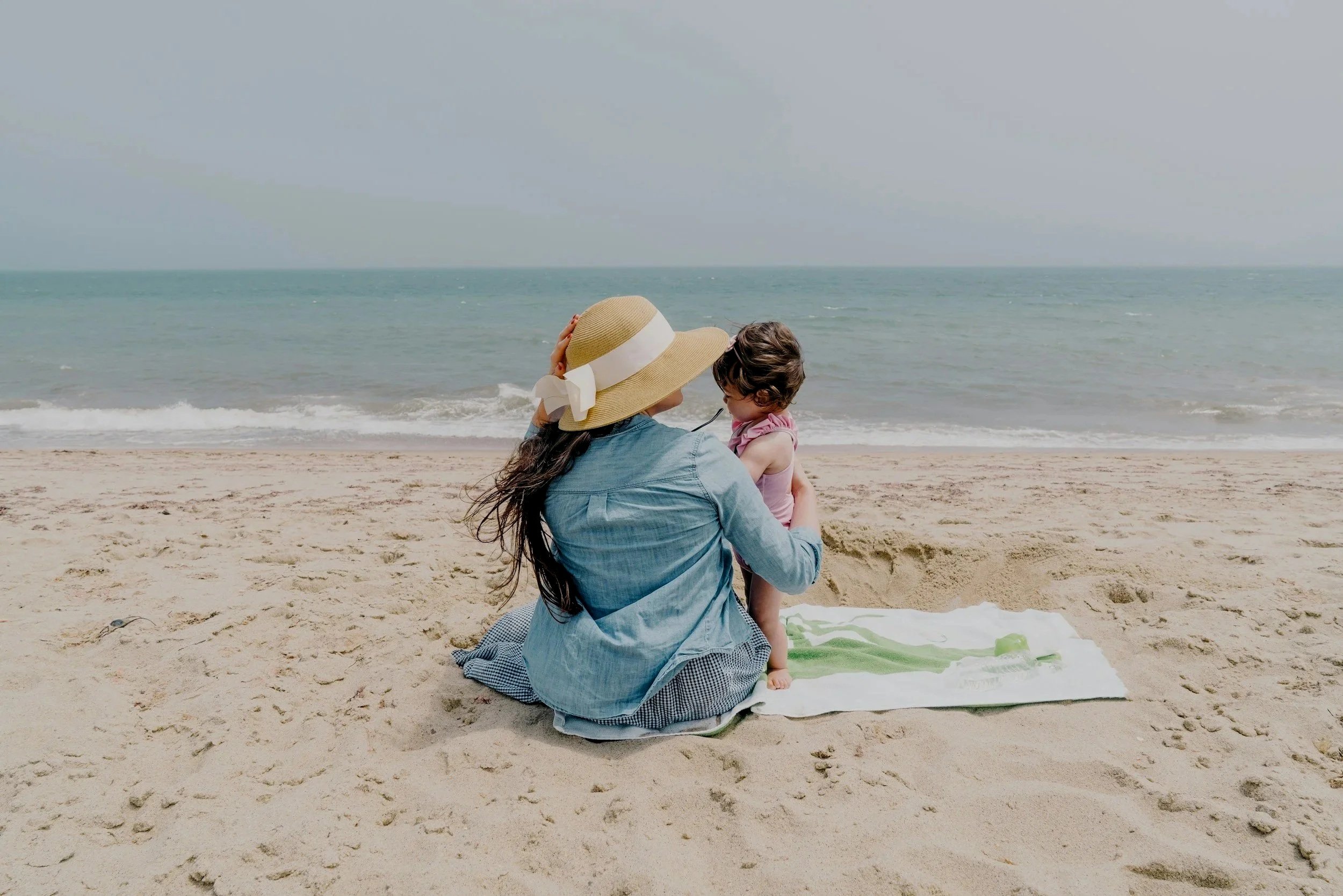 A woman and a young girl sitting on a beach towel facing the ocean, with the woman holding the girl close, both enjoying a sunny day at the beach.