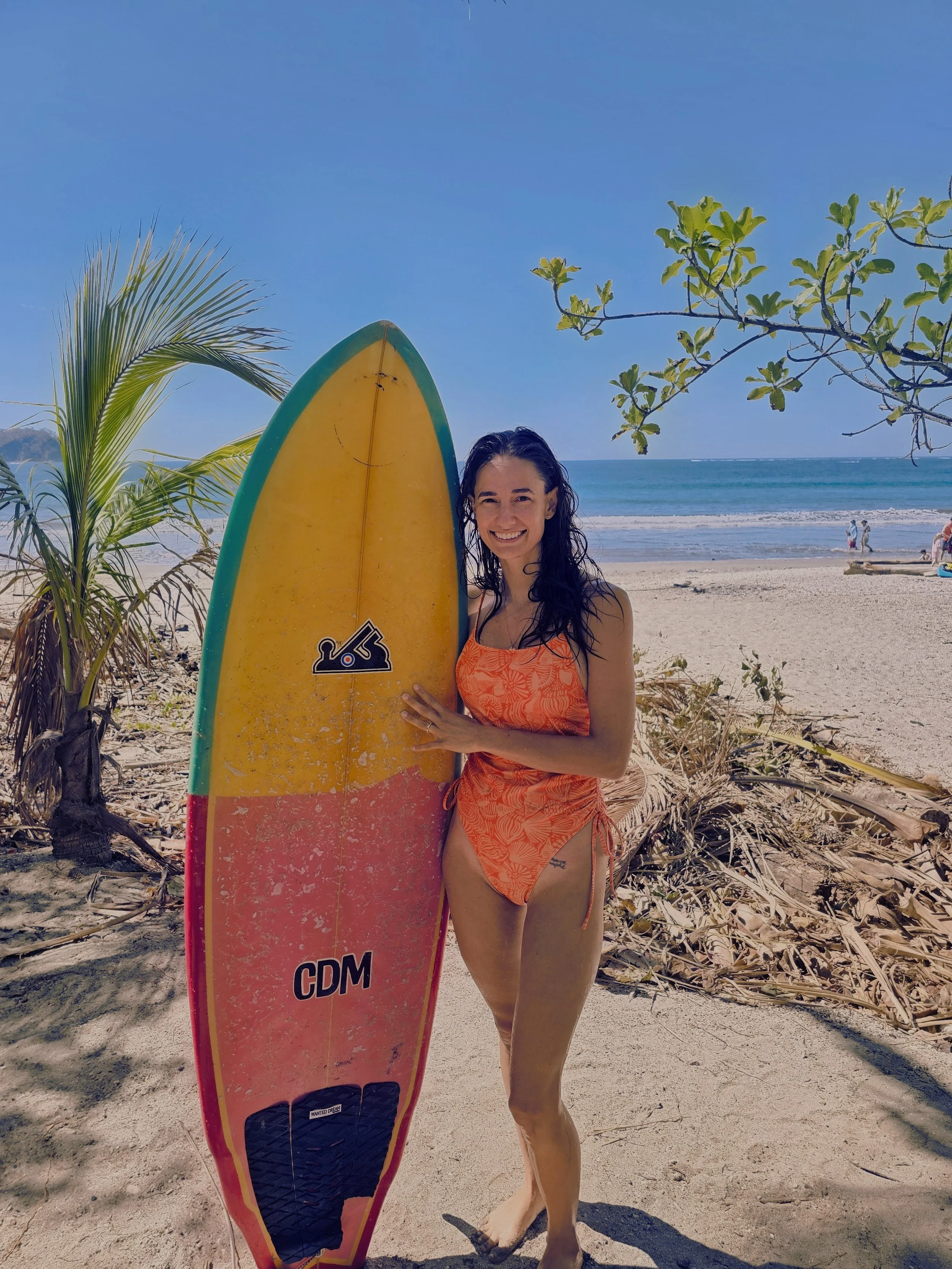 A woman in a red bikini standing on a sandy beach, holding a yellow and red surfboard, with the ocean and a clear blue sky in the background.