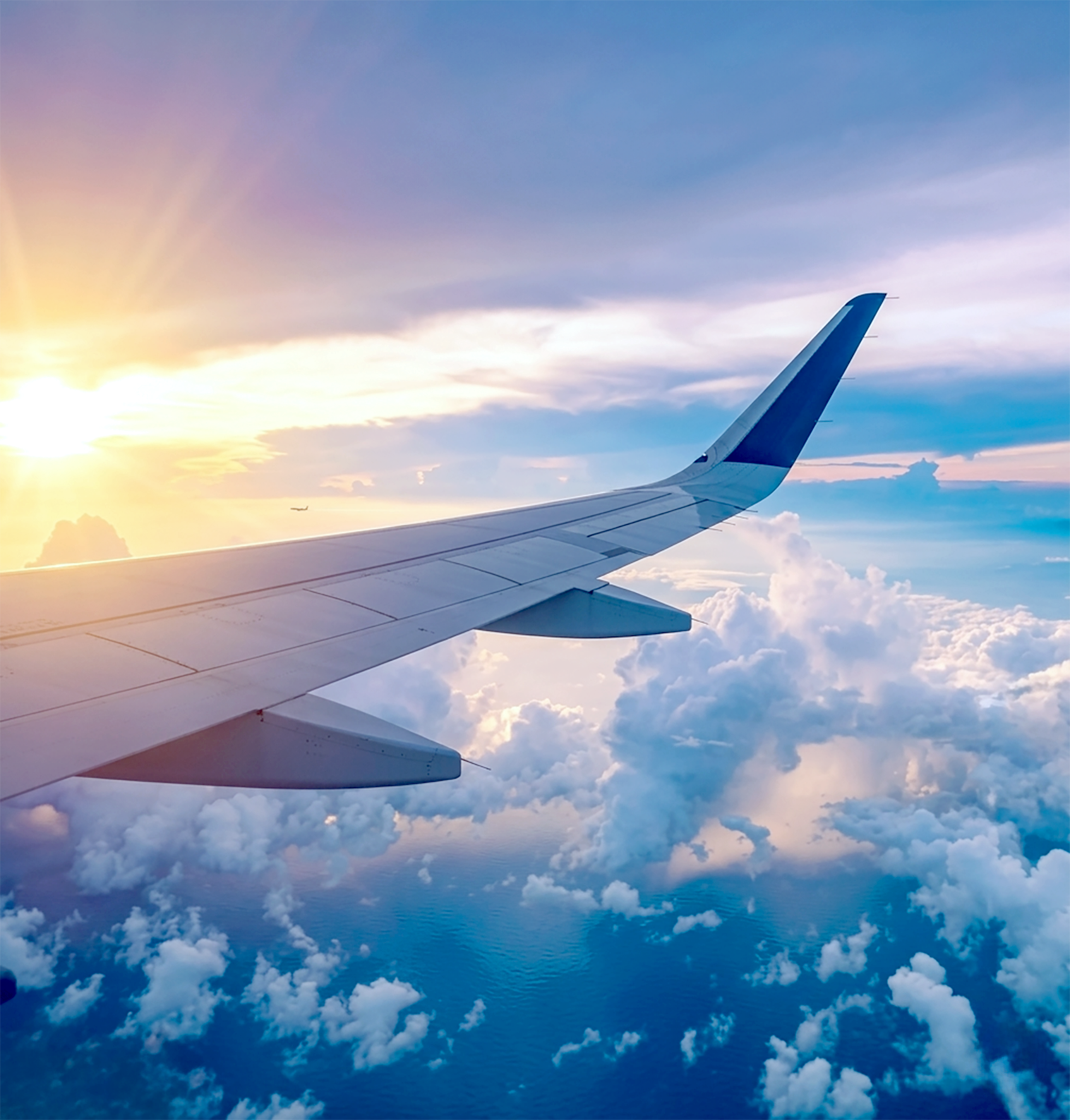 View from airplane window showing airplane wing, clouds, ocean, and sunset sky with a small airplane in the distance.