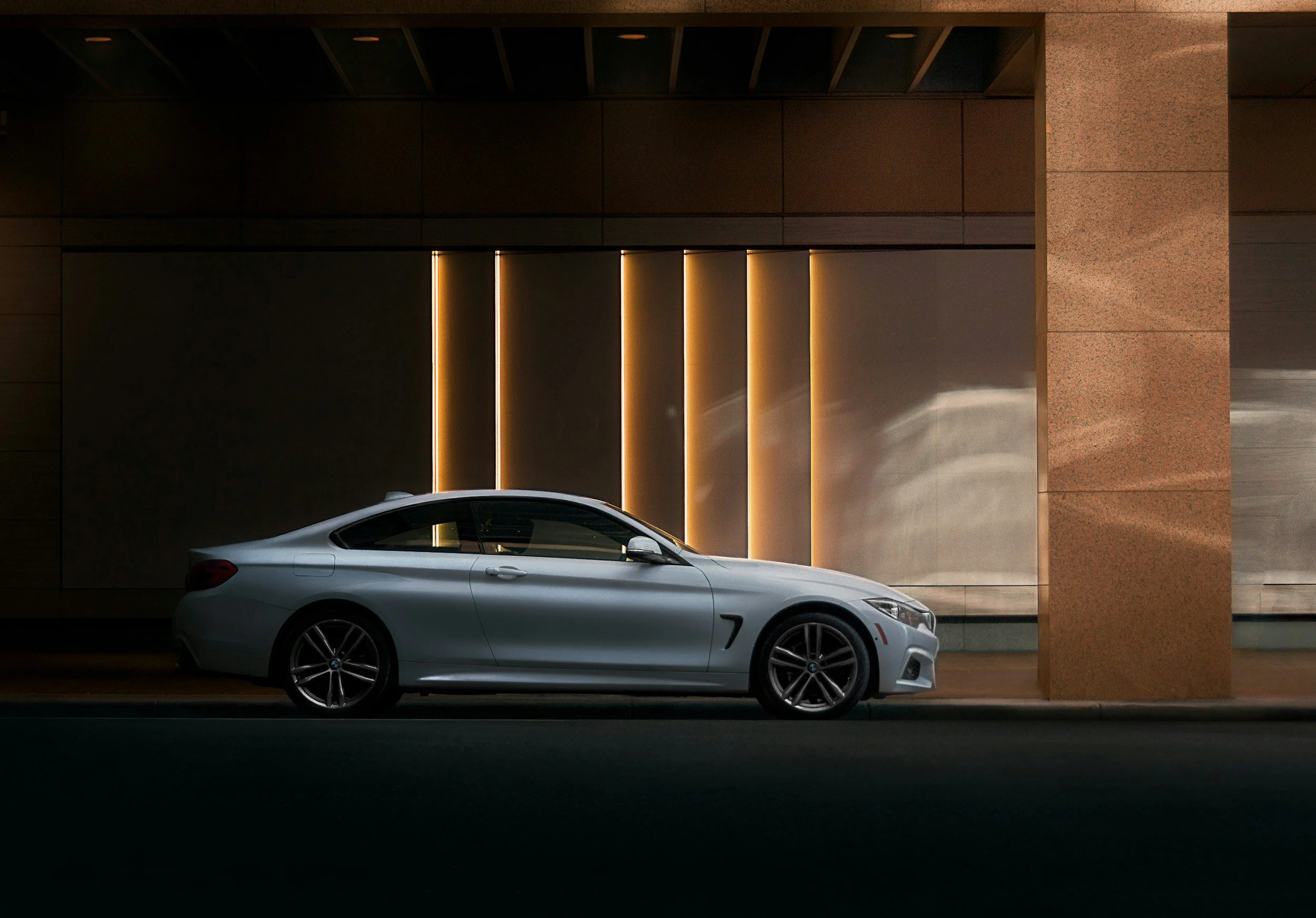 A silver luxury coupe car parked on the street against a modern building with illuminated vertical light accents.