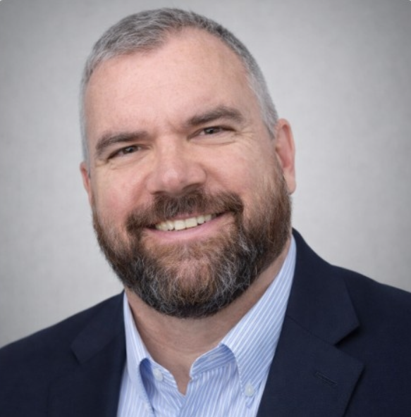 A smiling man with a beard and short gray hair, wearing a dark suit jacket and a light blue dress shirt, against a plain gray background.