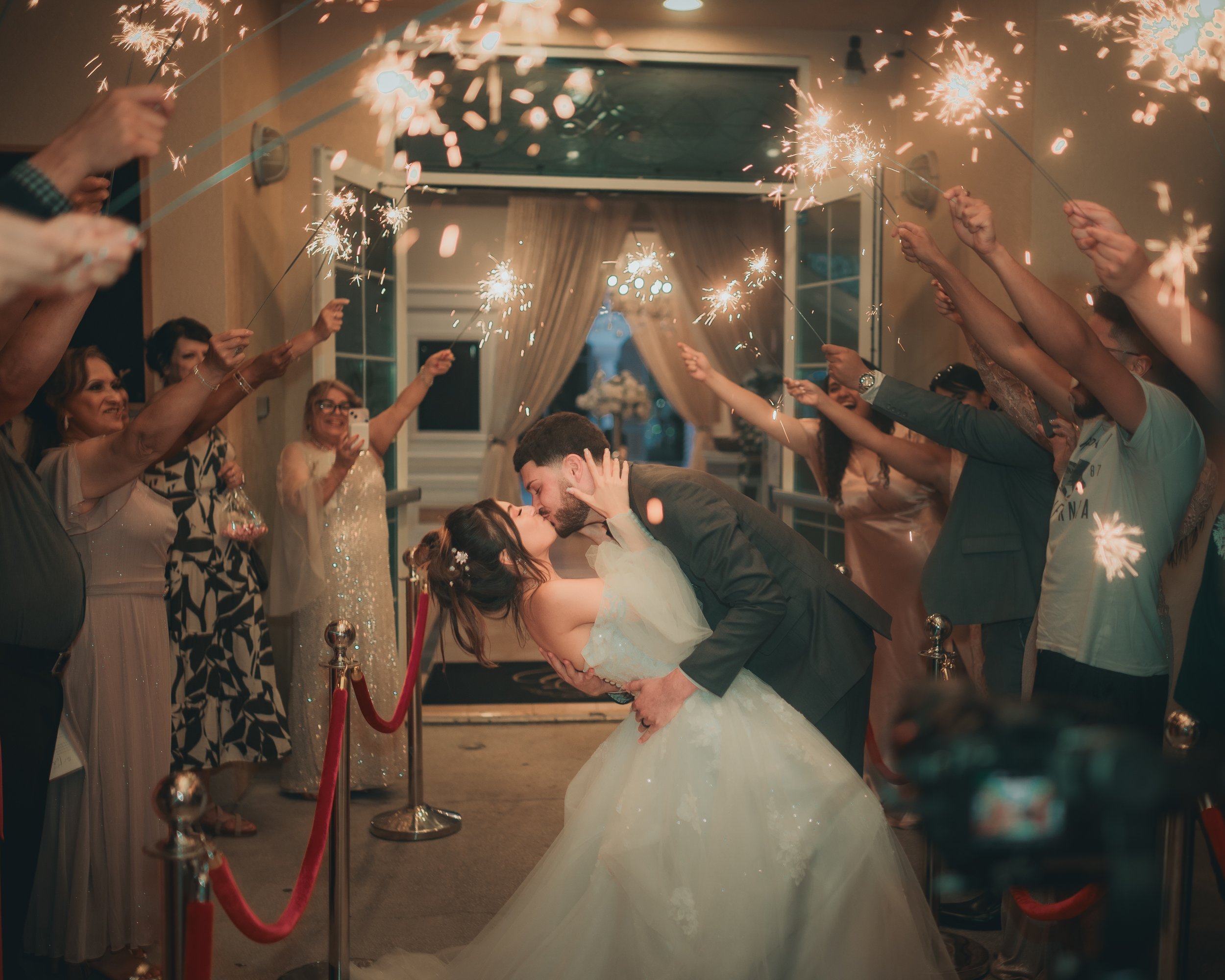 A newlywed couple kisses amid friends holding sparklers at night outside a wedding venue.