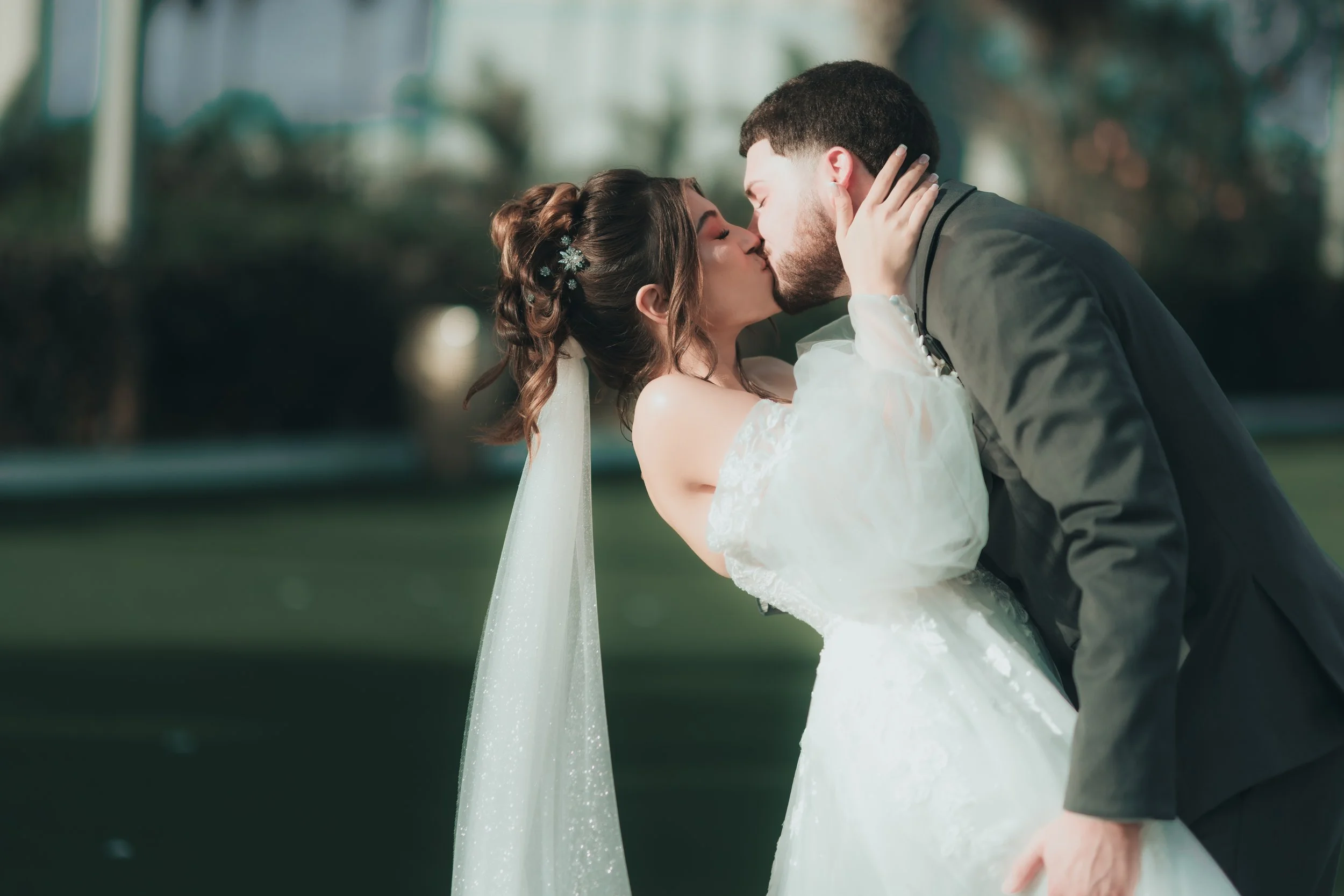 A bride and groom sharing a kiss outdoors on their wedding day, with the bride wearing a white wedding dress and the groom in a dark suit.