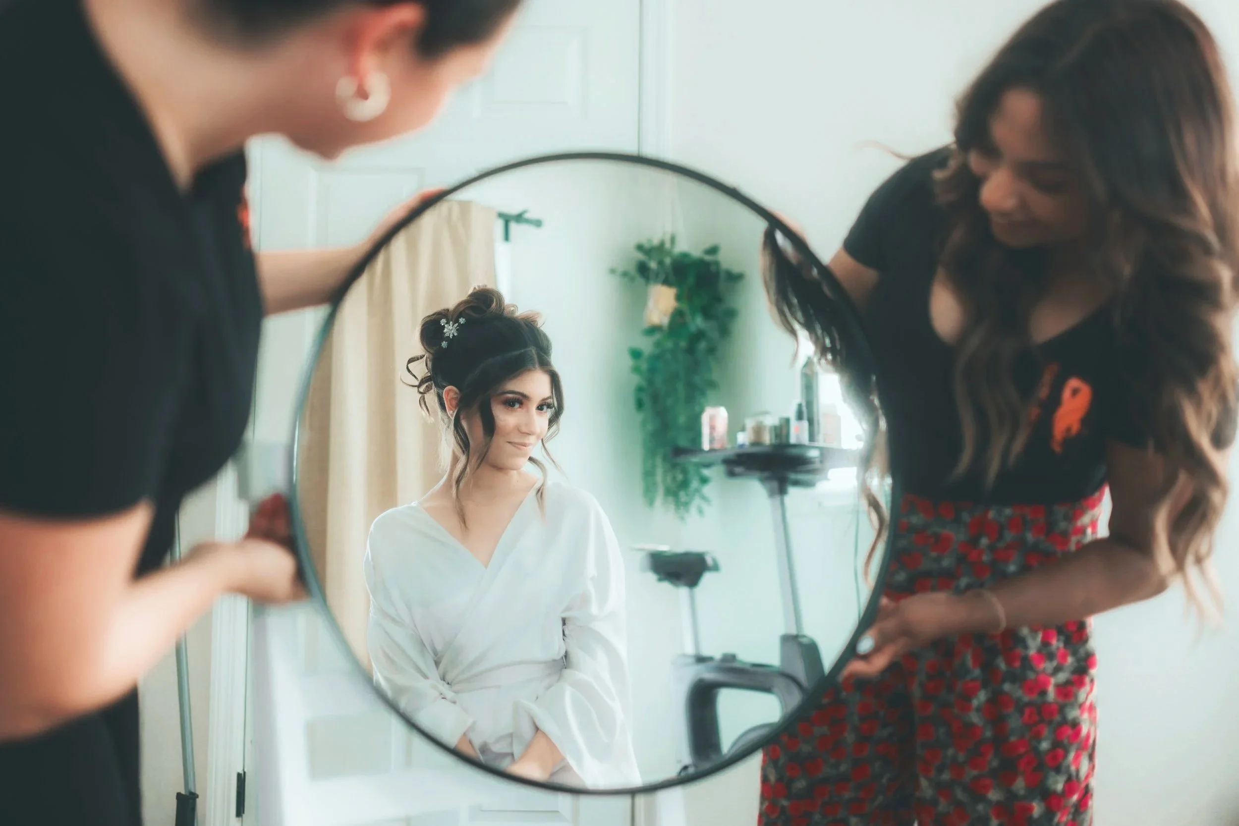 A woman in a white robe sits in front of a mirror, while another woman with long wavy hair, wearing a black top and red patterned pants, styles her hair.