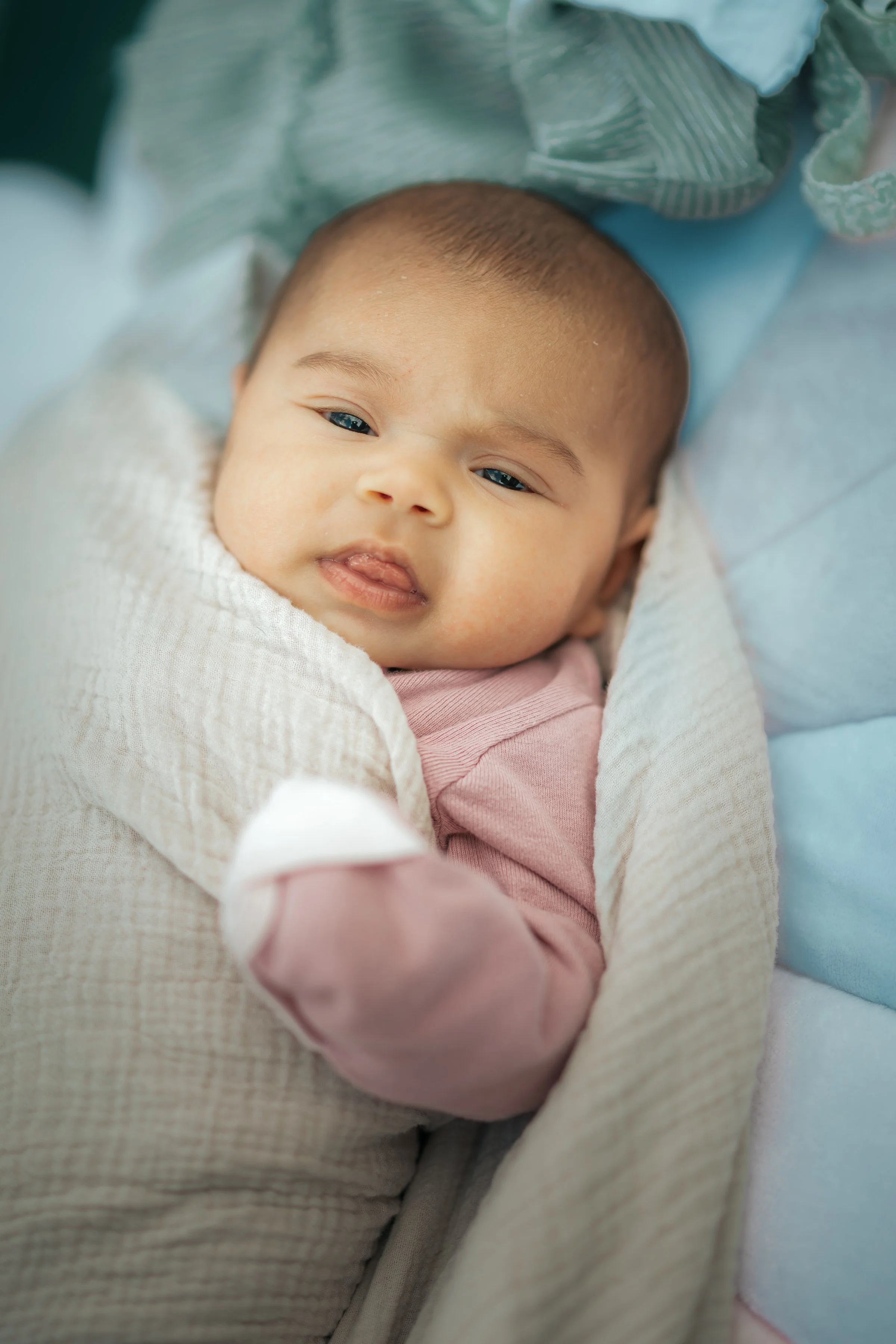 A baby lying on a cushioned surface, wrapped in a beige blanket, wearing a pink long-sleeve shirt, with short dark hair and light skin, looking at the camera.