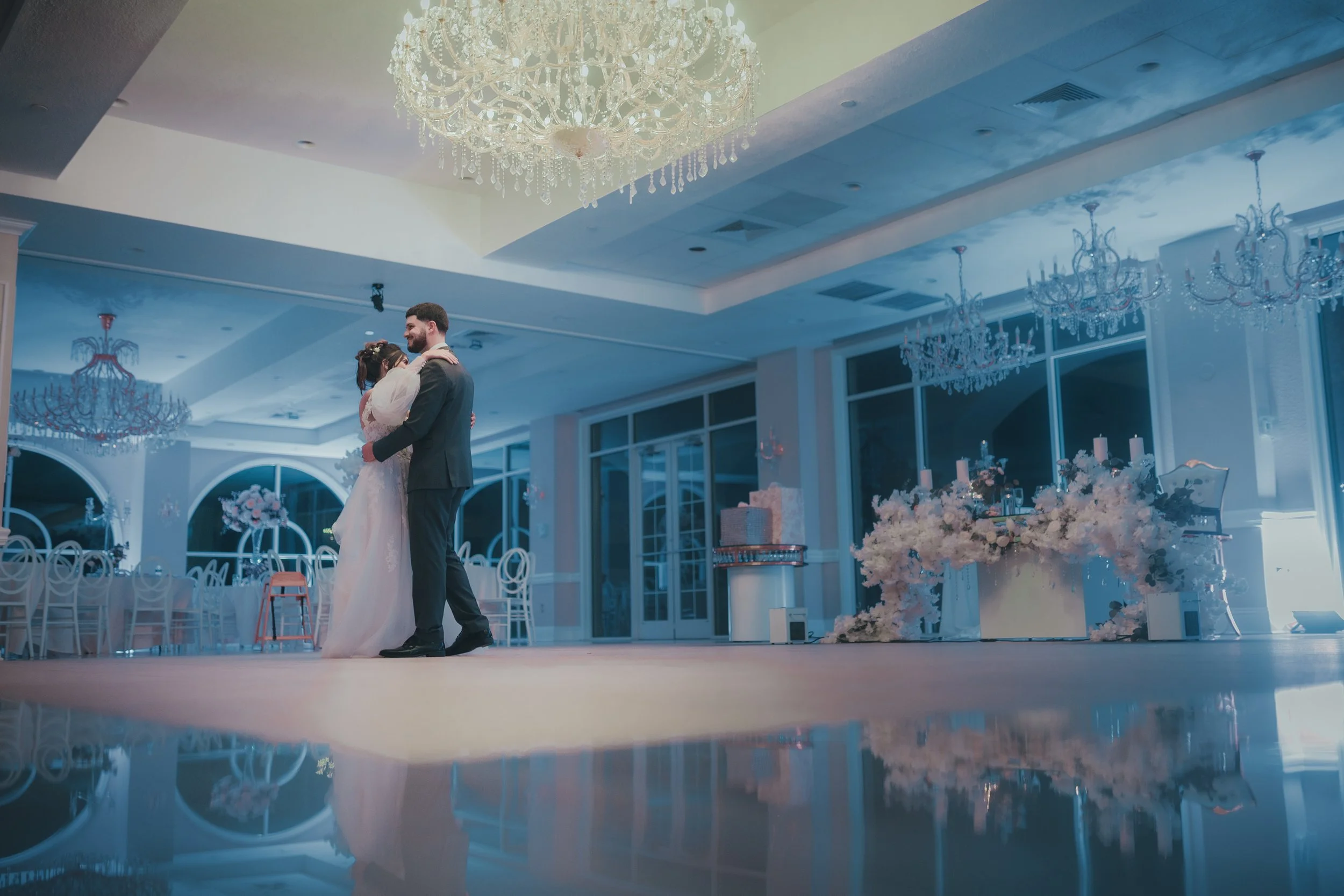 A bride and groom dancing in a wedding venue decorated with chandeliers, floral arrangements, and elegant lighting.