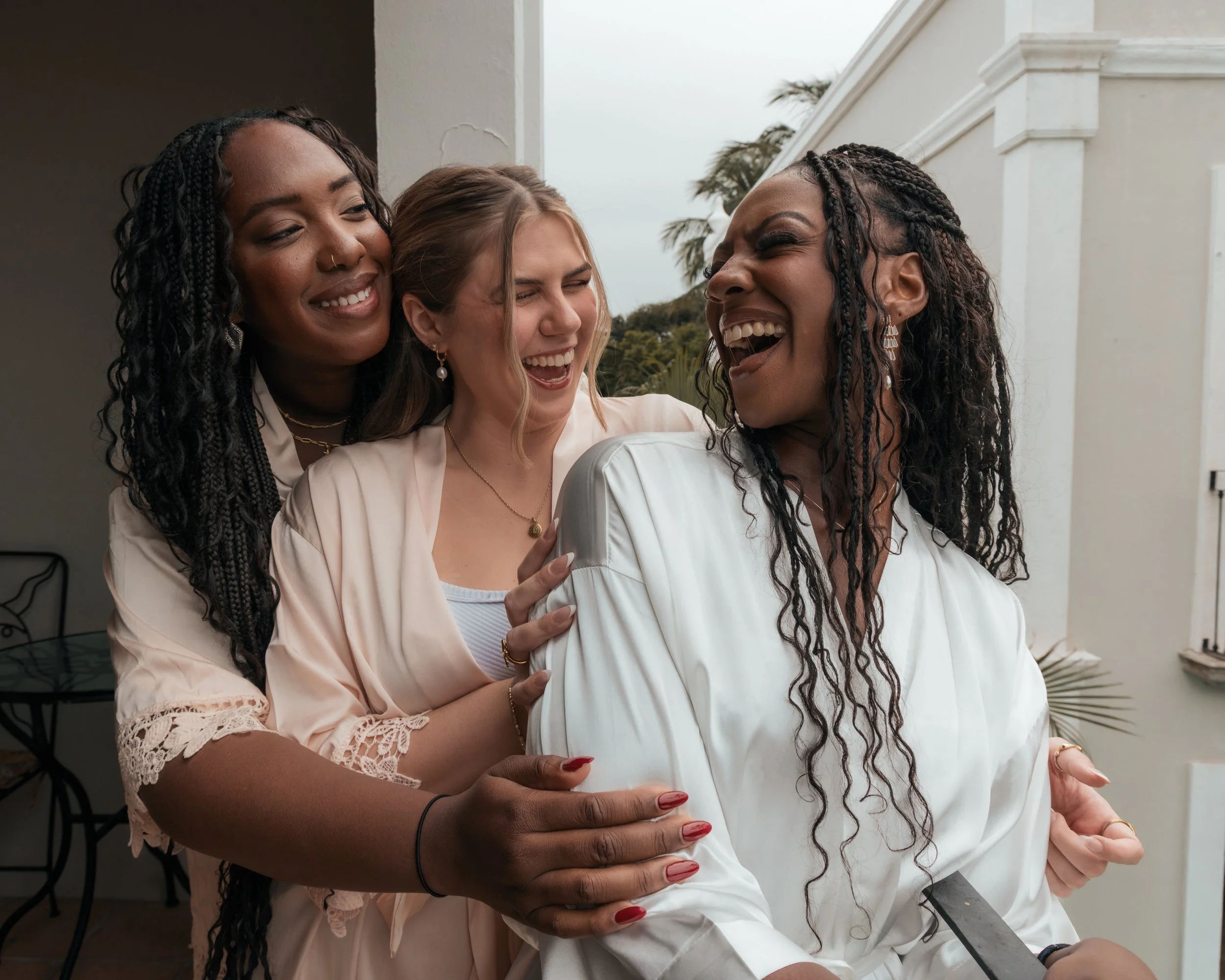 Three women laughing and hugging on a balcony, dressed in white or light-colored clothes.