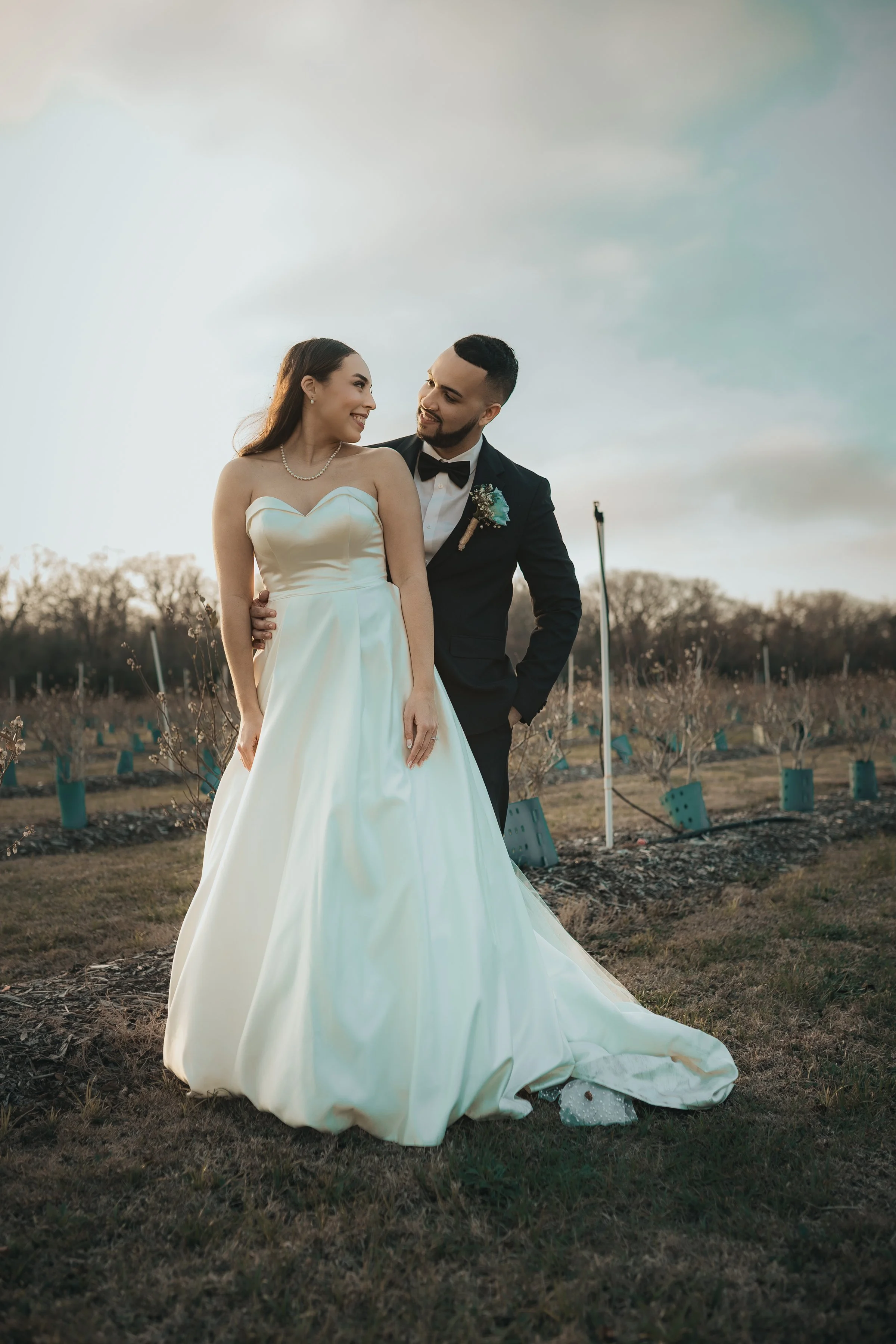 A bride and groom stand together in a field during sunset, smiling at each other. The bride wears a strapless white wedding gown with a pearl necklace, and the groom wears a black tuxedo with a bow tie and boutonnière.