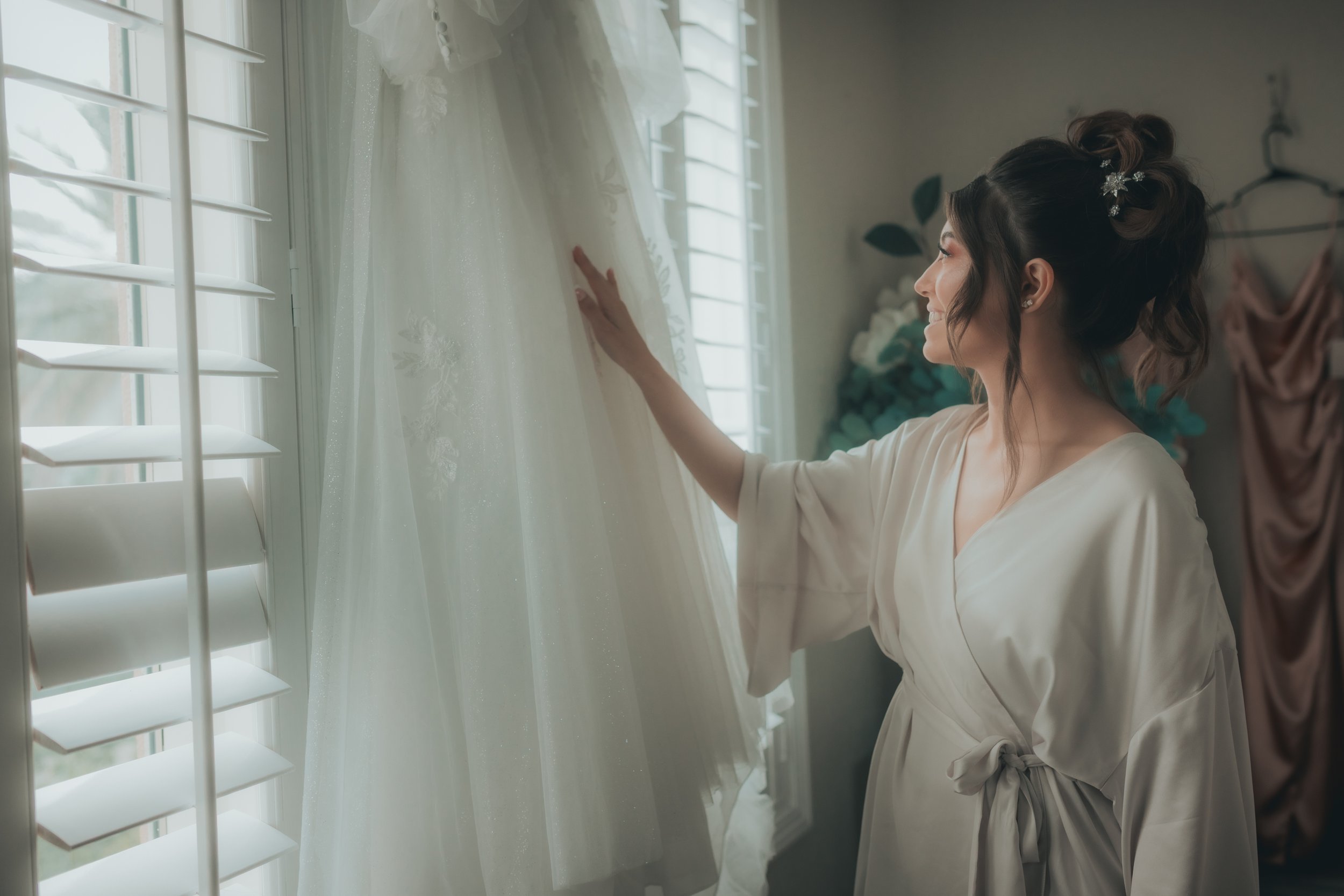 A woman in a silk robe touching a wedding dress hanging by a window.
