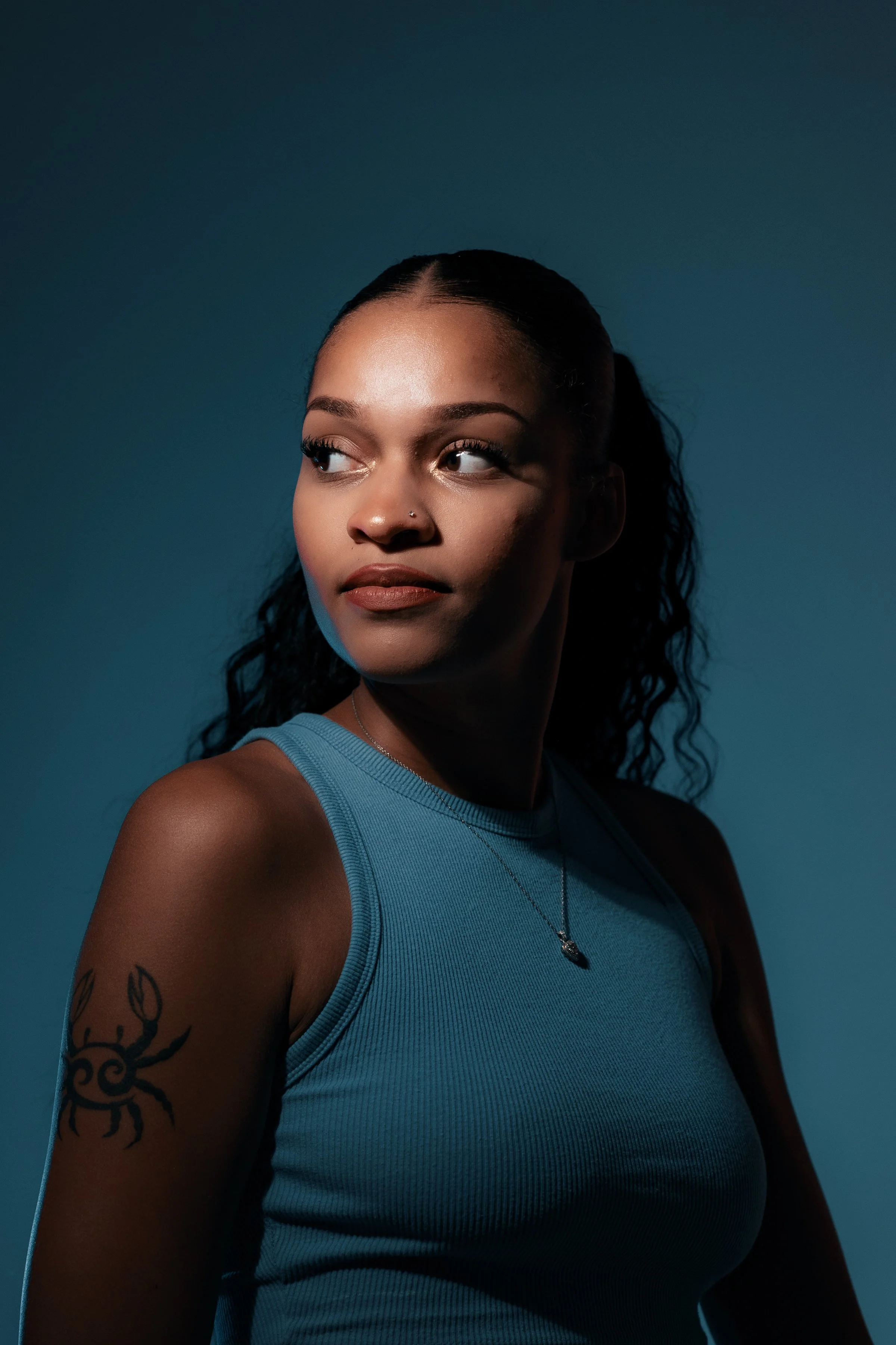 Portrait of a young woman with dark curly hair, tan skin, wearing a blue sleeveless top and a necklace, against a dark blue background.