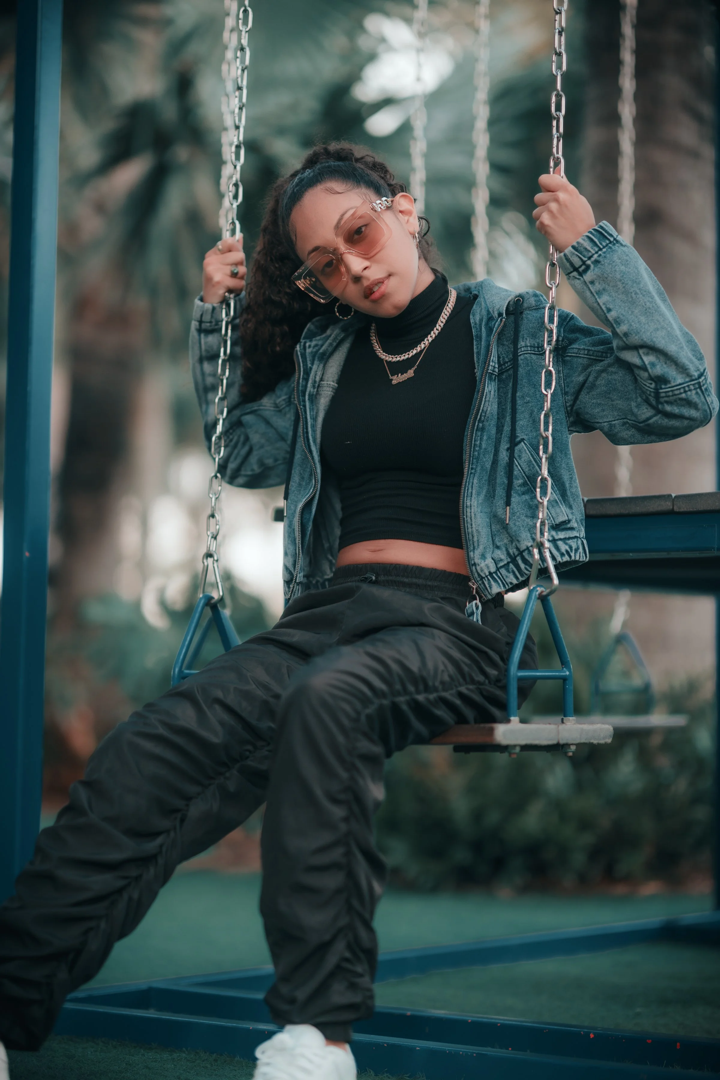 A young woman with curly hair, wearing sunglasses, a black crop top, a denim jacket, black pants, and white sneakers, sitting on a swing outdoors with trees in the background.
