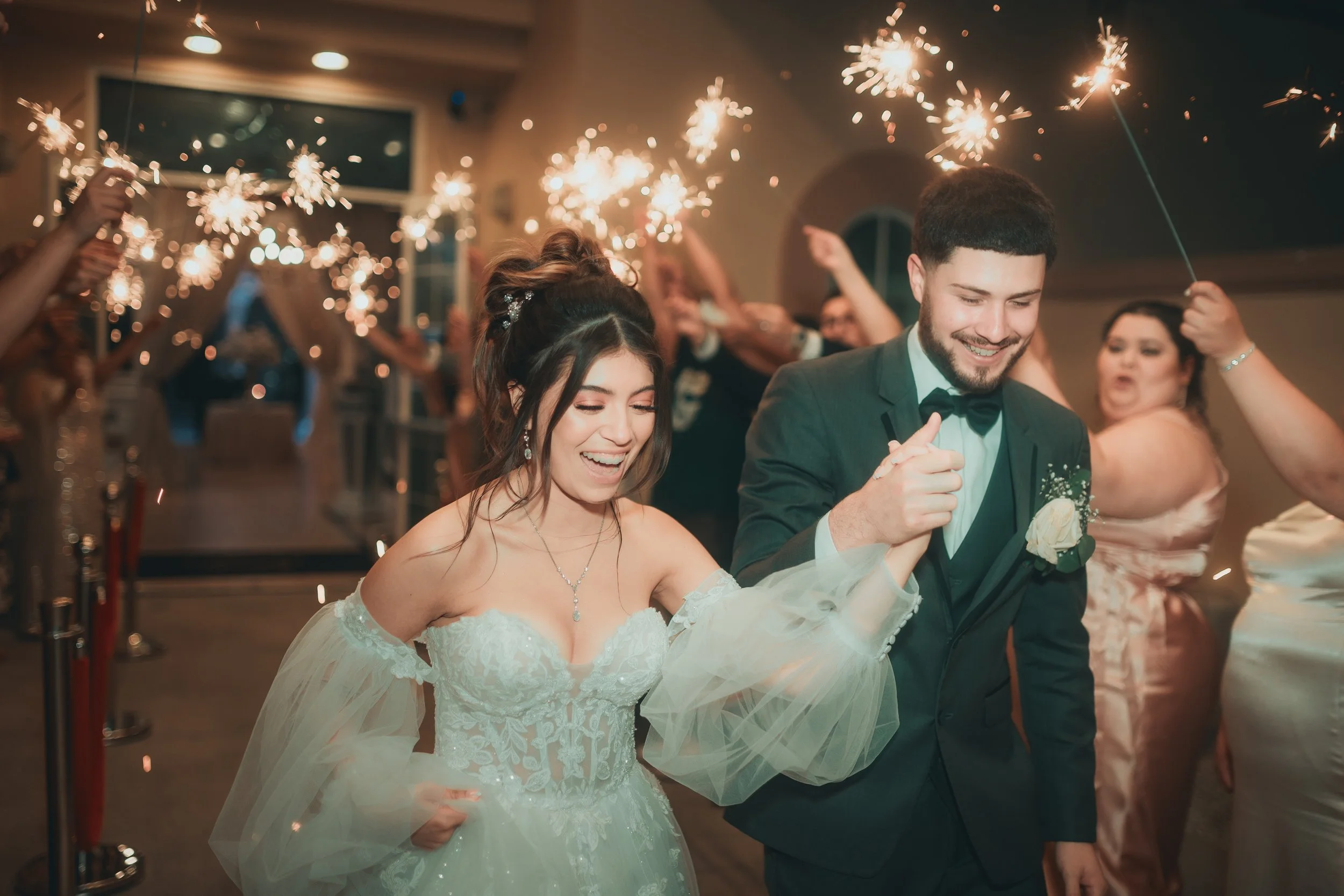 A bride and groom holding hands and smiling at their wedding reception, with sparklers and guests celebrating in the background.
