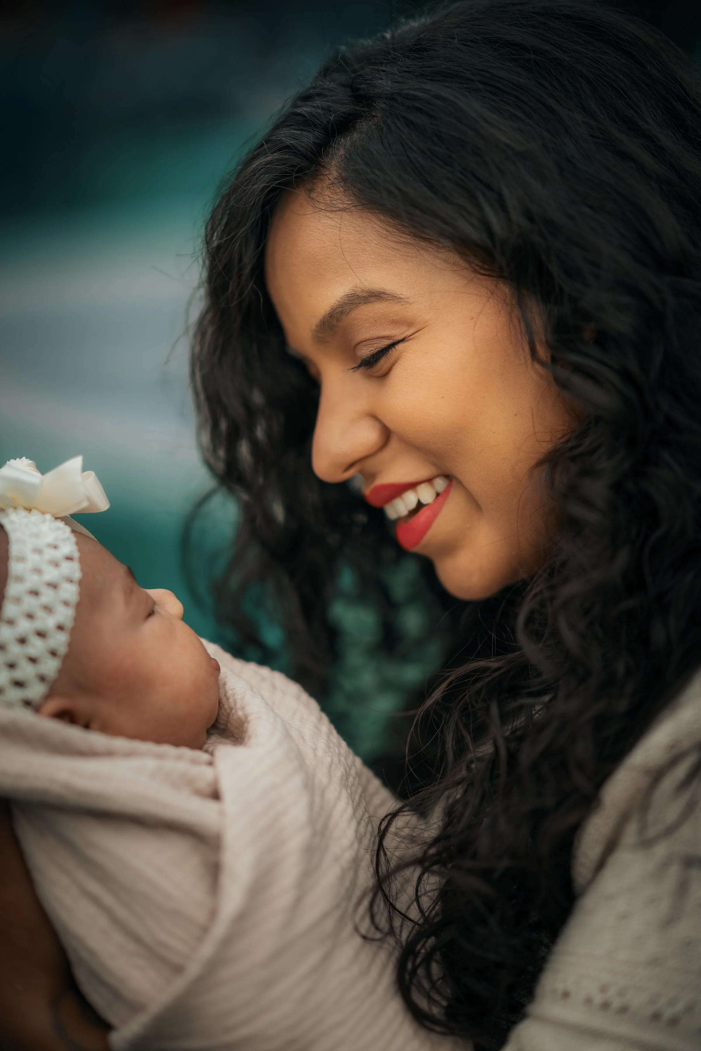 A woman with long dark curly hair smiling and holding a newborn baby wrapped in a blanket, both facing each other in a close-up shot.